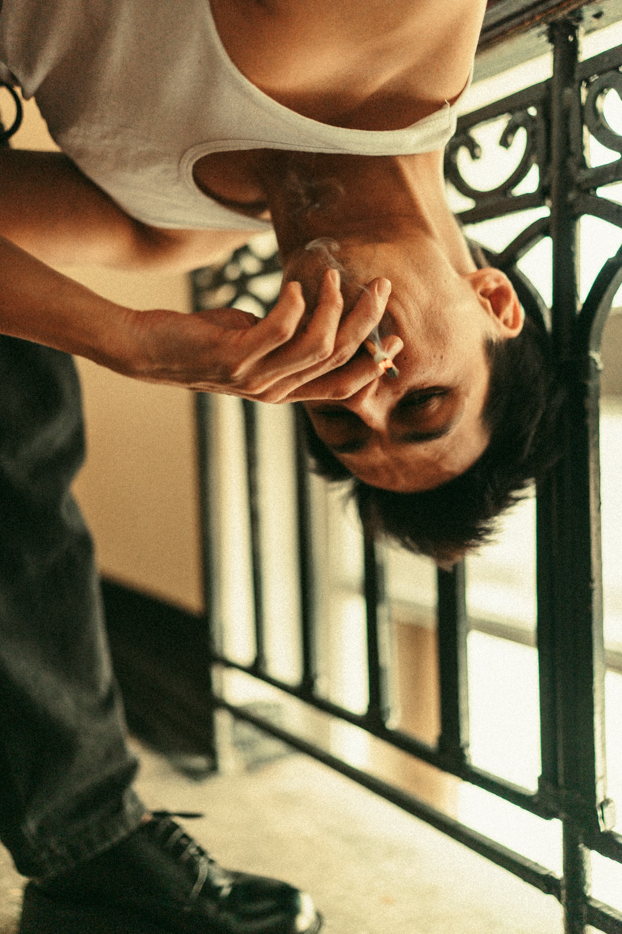 Мужчина в белой майке с сигаретой сидит на лестничной клетке в старом здании, опираясь на перила, создавая атмосферу уличной жизни