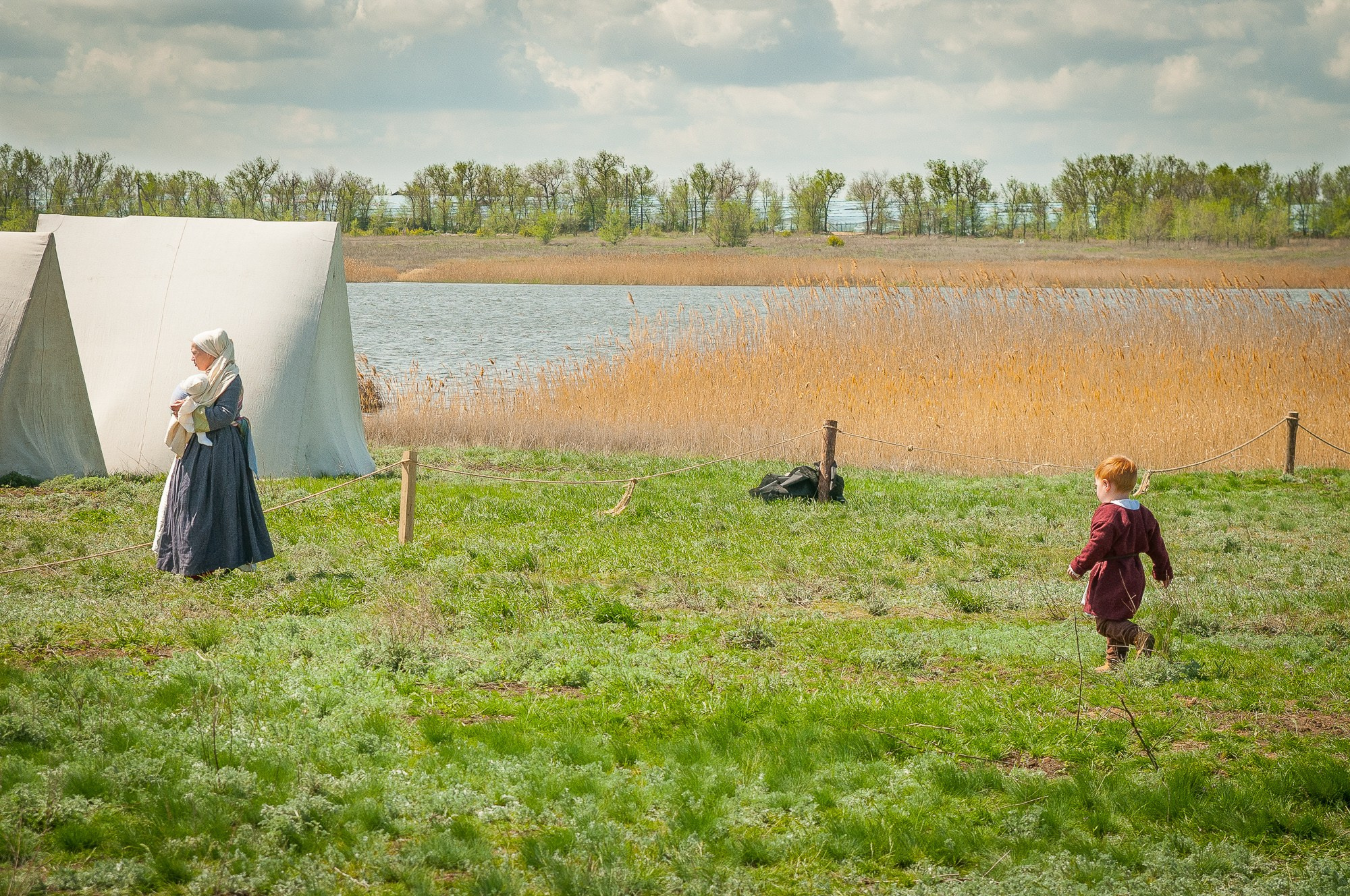 Маленький мальчик в историческом костюме идёт по полю с травой на берегу озера