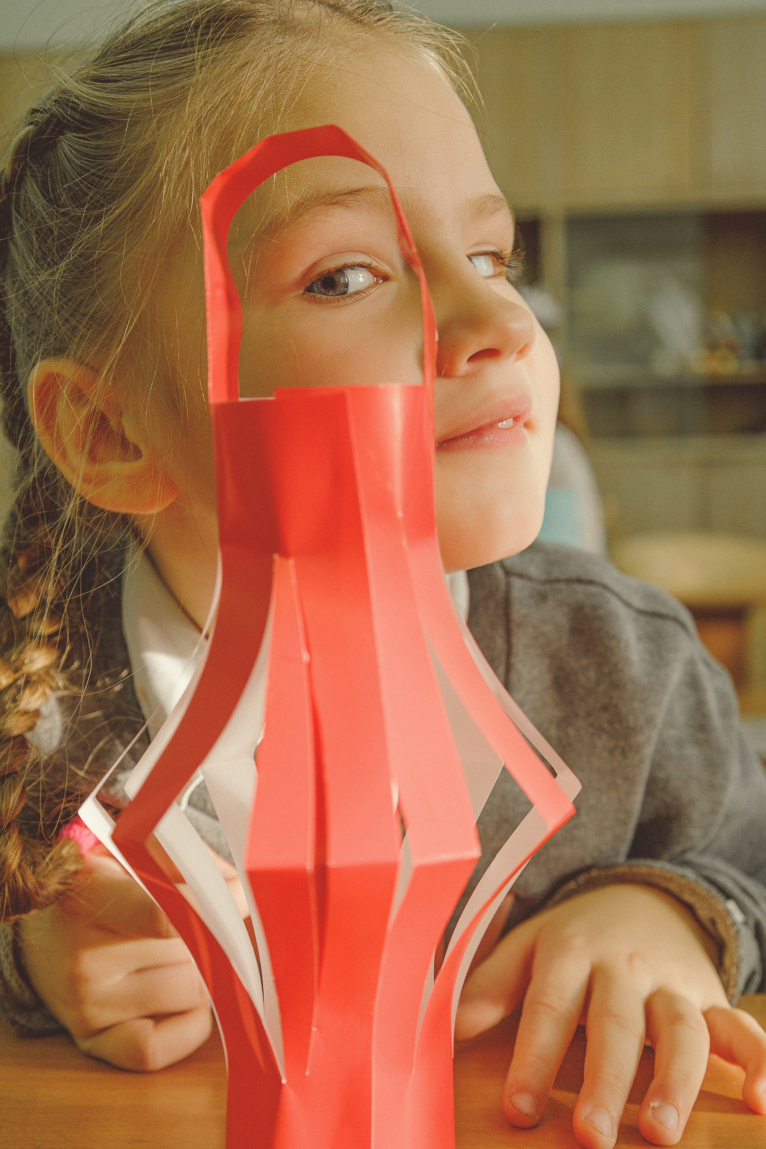 Девочка с заплетенными в косу волосами и игривым выражением лица выглядывает из-за красного бумажного фонаря, который она держит на столе, в тепло освещеннойм школьном классе