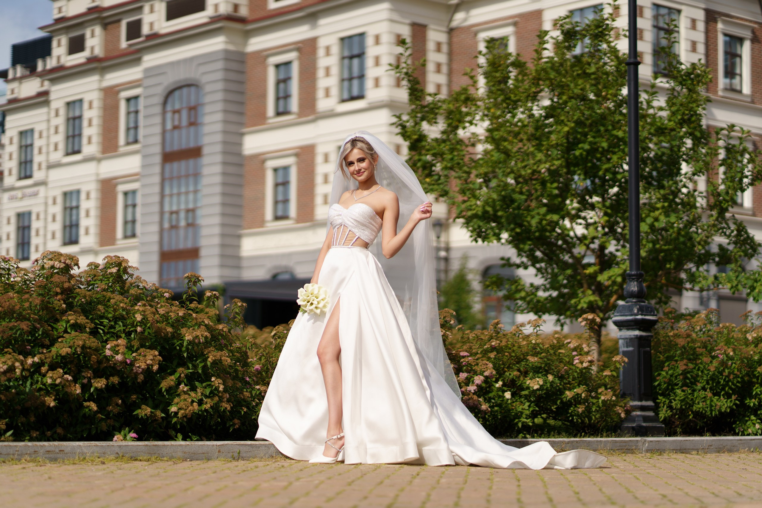 Невеста в современном свадебном платье с разрезом позирует на фоне архитектурного здания. Её образ дополняют фата, белые туфли и букет свежих цветов. Вокруг — зелёные кусты и деревья | свадебная съемка в общественных местах Екатеринбург