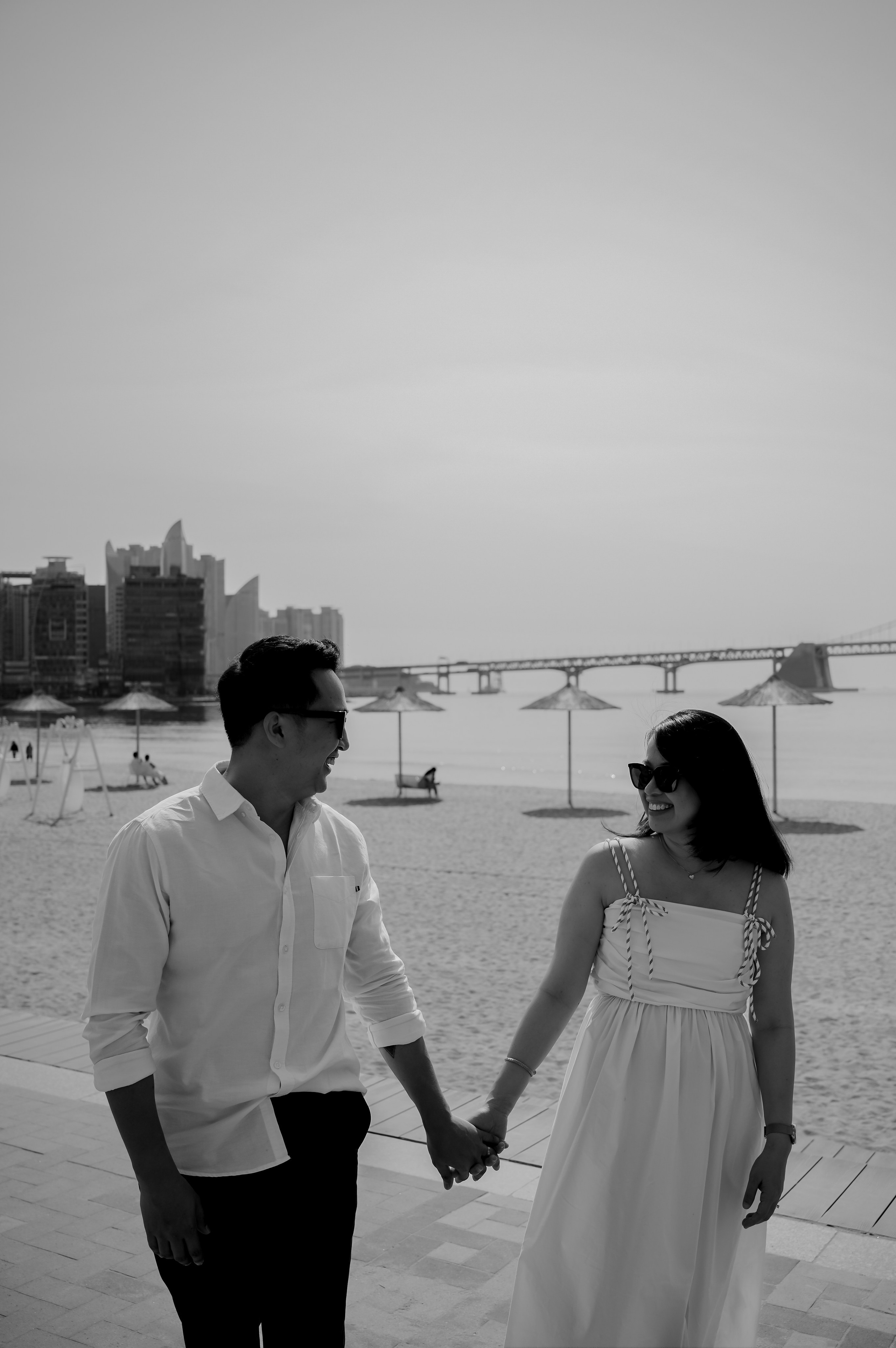 Couple holding hands on Gwangalli Beach with a view of Gwangan Bridge, Busan — black and white photo · 부산 여행 스냅