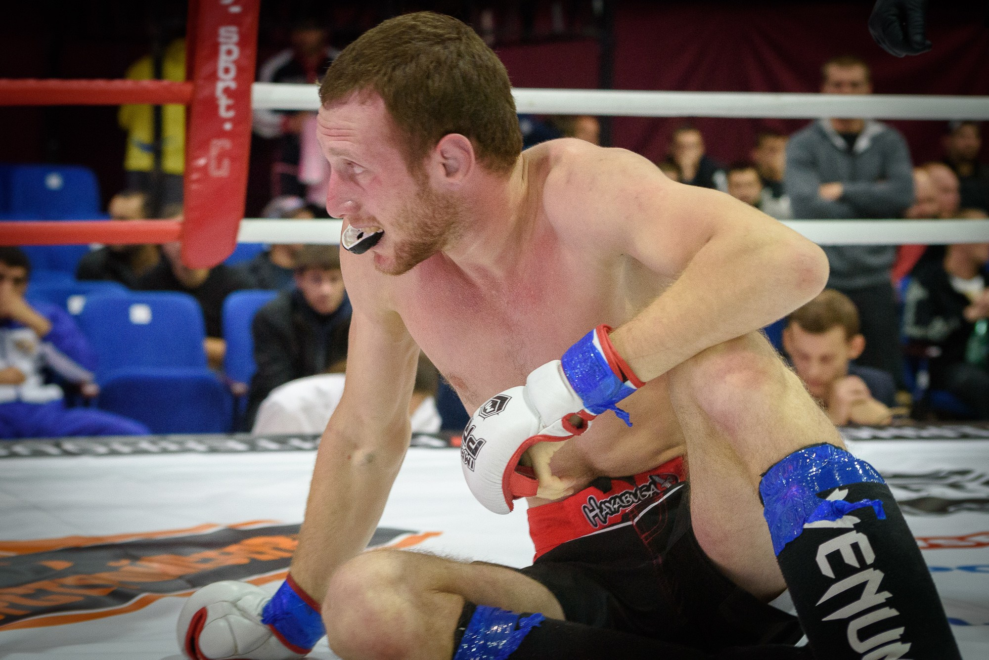 Боец сидя на ринге смотрит в сторону соперника, высунув капу изо рта