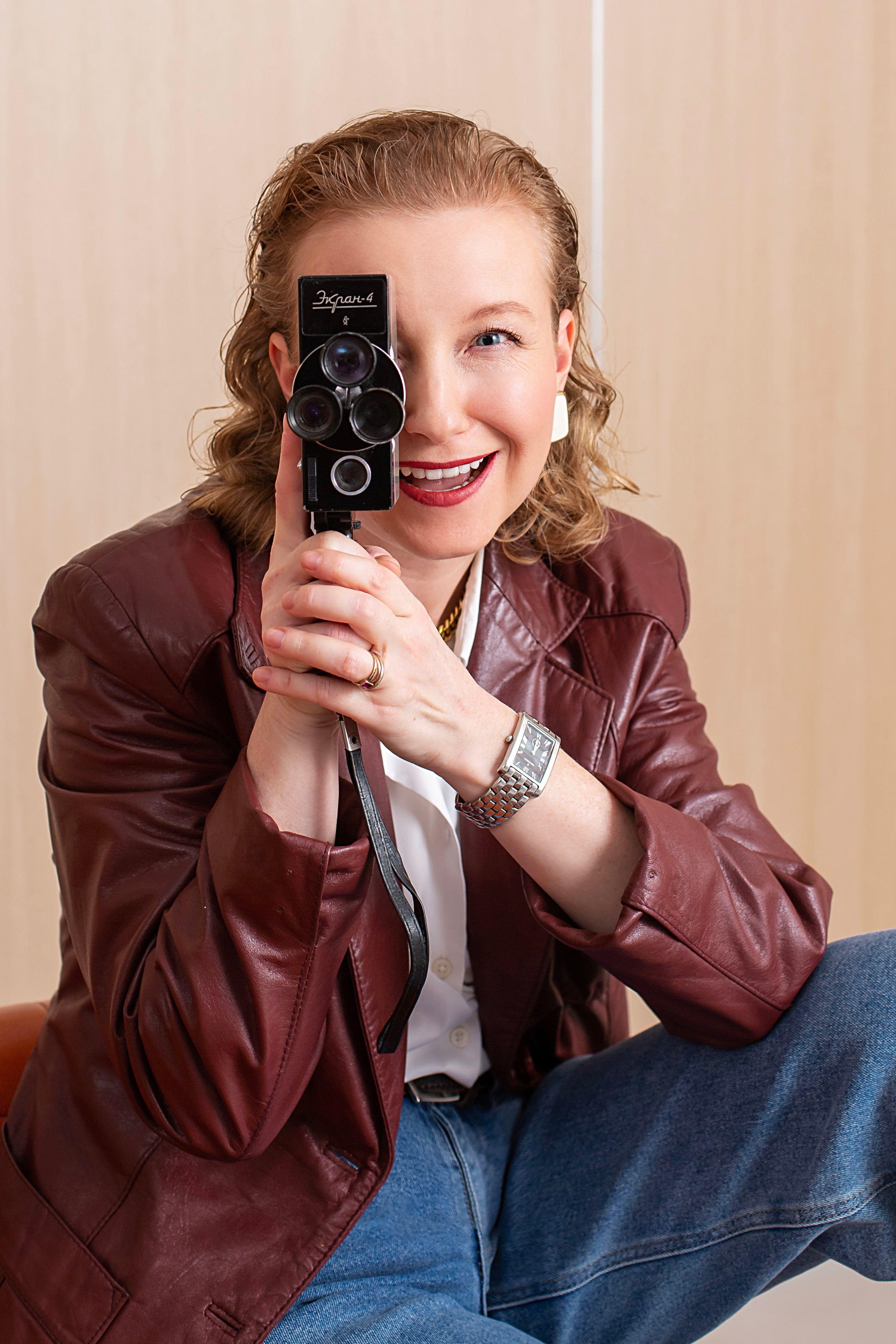 Фотосъемка в ретро стиле, женщина в стильном малиновом кожаном пиджаке изображает оператора с видеокамерой в руках