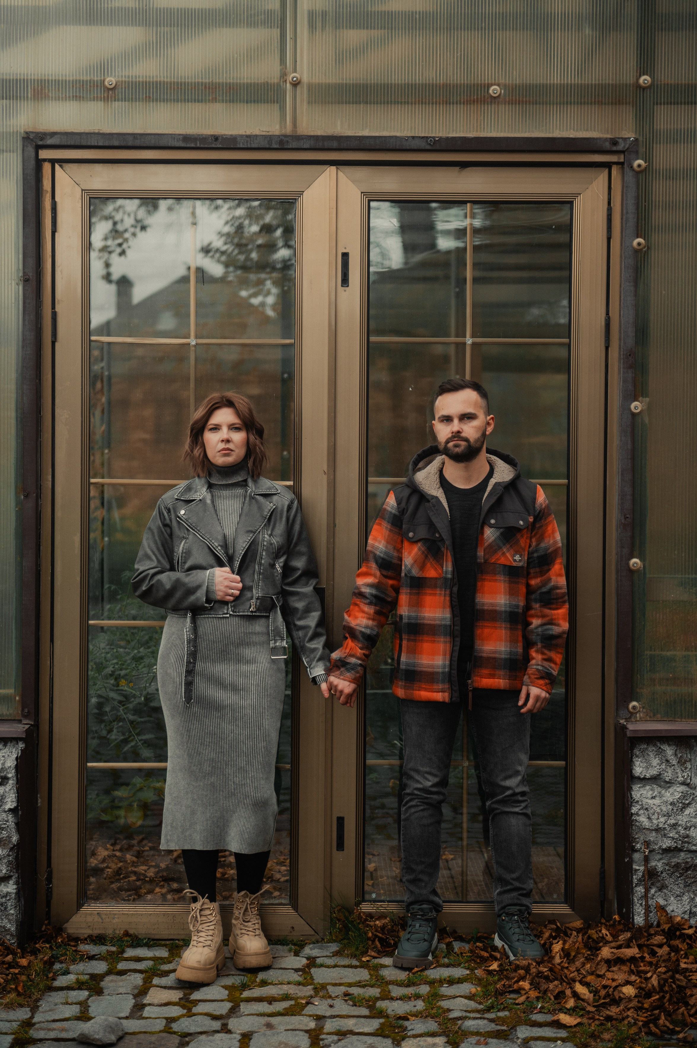 Парень в клетчатой куртке и девушка в сером платье держатся за руки у стеклянных дверей. Вокруг опавшие листья, атмосфера спокойная.