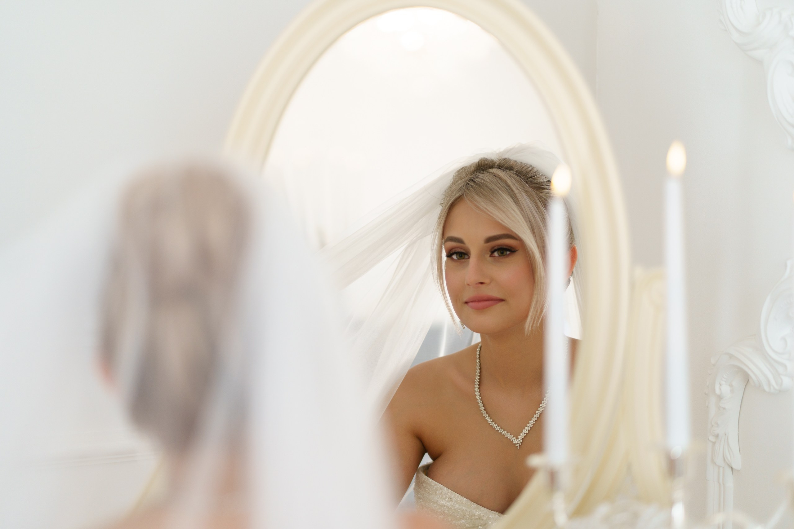 Невеста в белом платье и фате рассматривает своё отражение. Жемчужное колье изящно дополняет её образ. На переднем плане — свечи, создающие тёплый свет | профессиональная свадебная фотосессия Екатеринбург