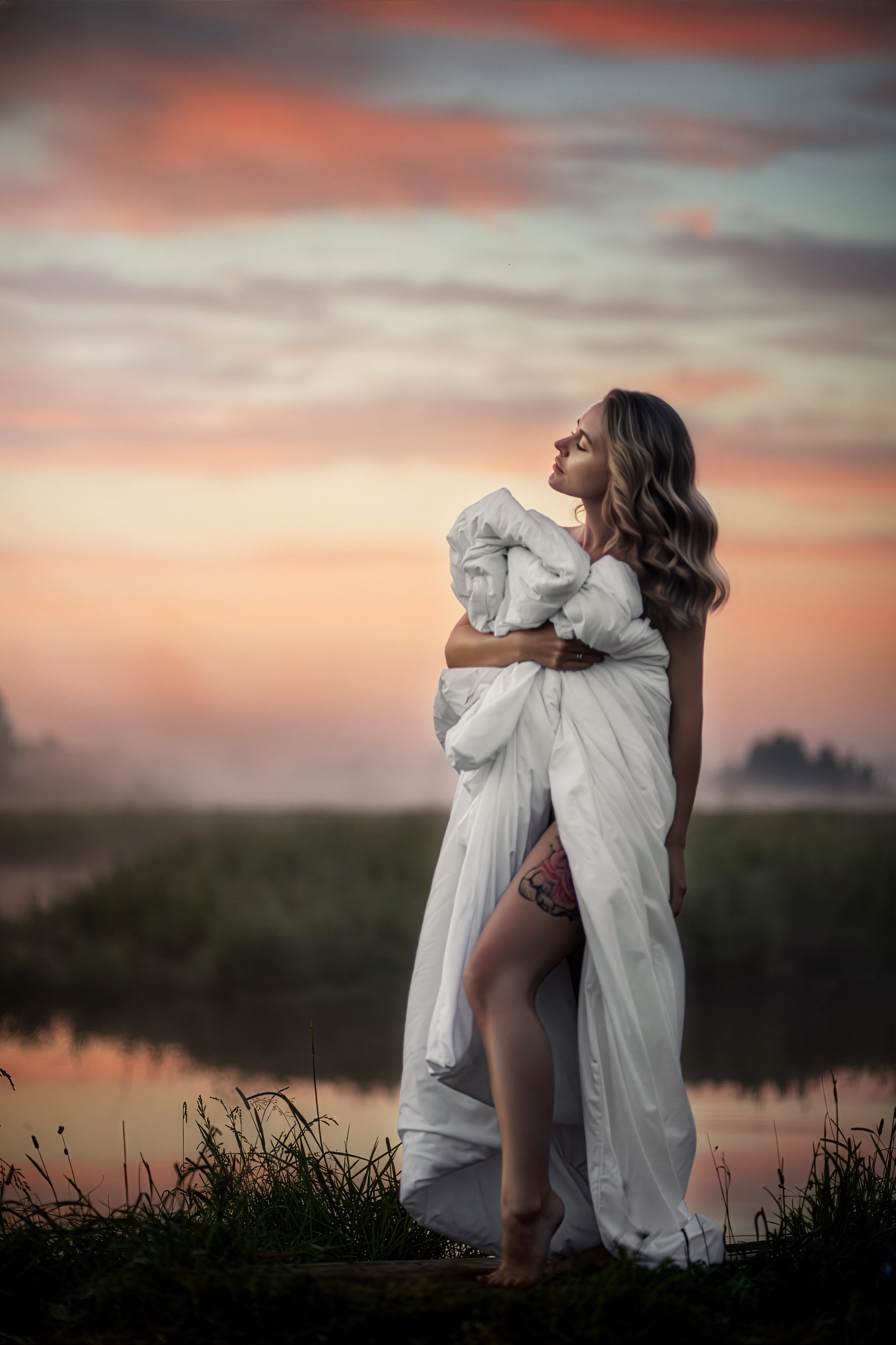 Женщина на рассвете у реки, на фоне тумана и персикового неба.