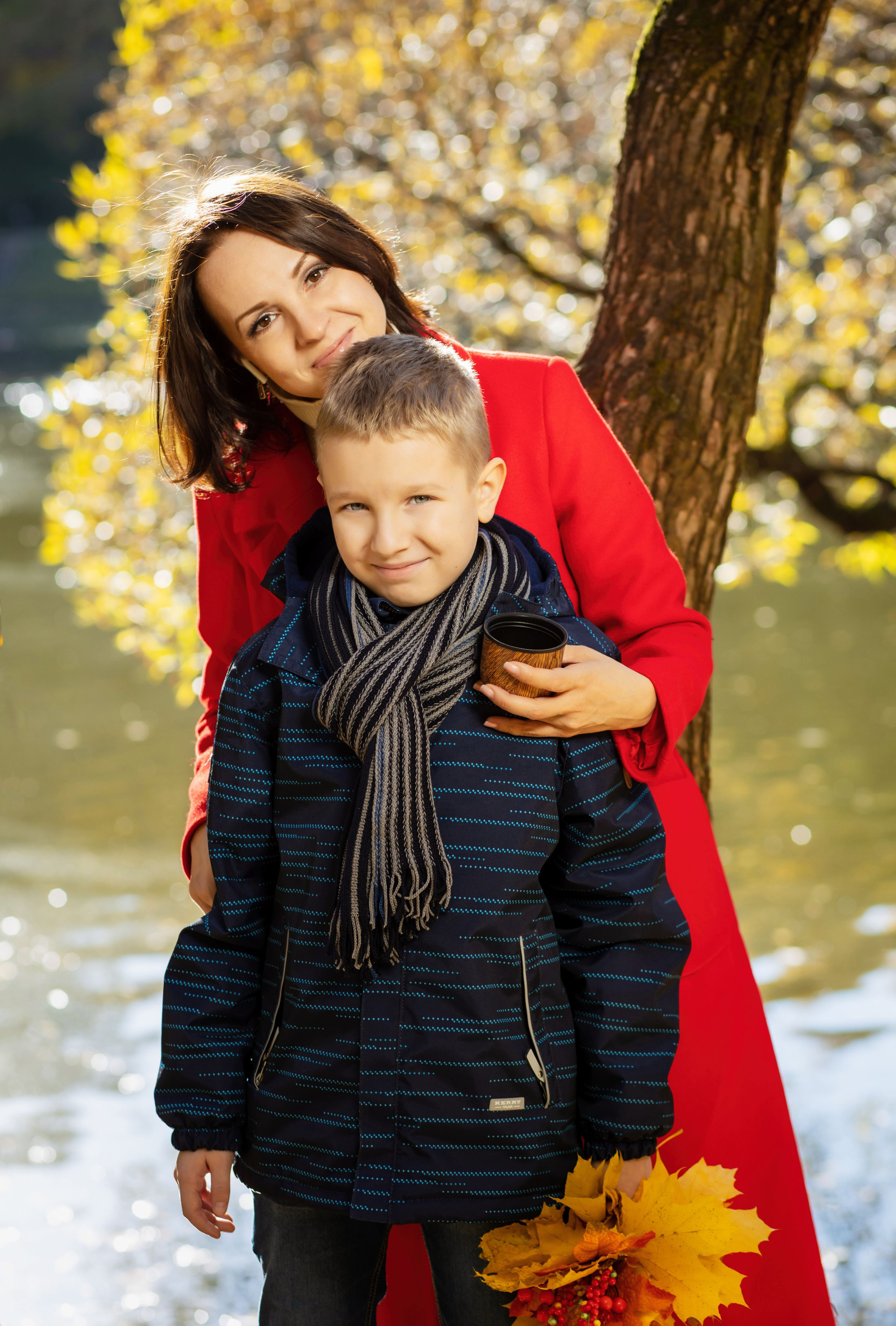 Мама в красном пальто обнимает сына на фоне осеннего дерева