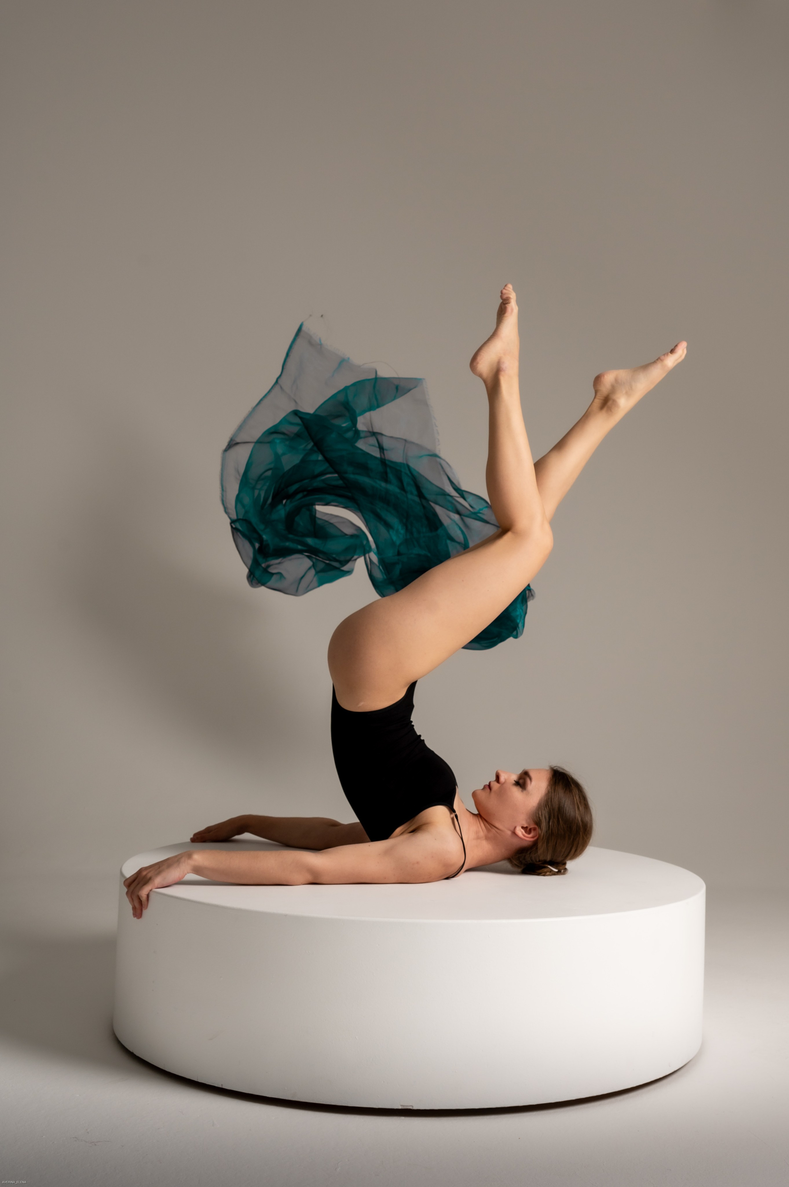 девушка гимнастка в поднятыми вверх ногами с летящей тканью на белом подиуме, фотосессия в студии