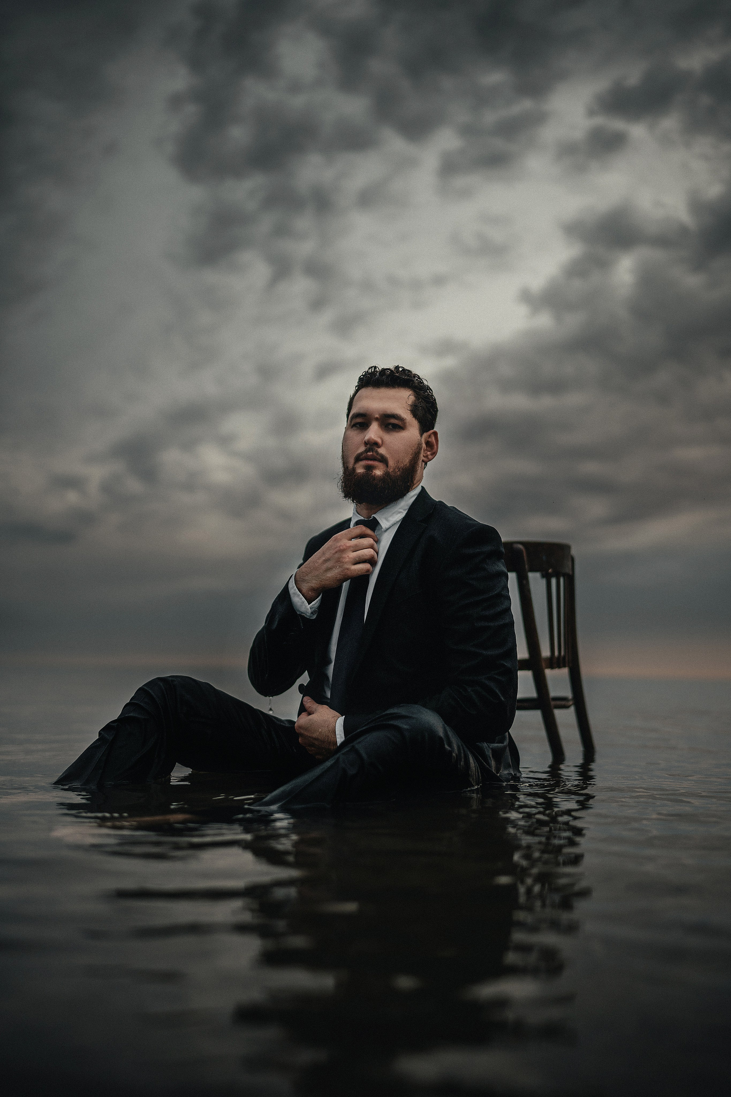 Мужчина в костюме сидит на стуле, погружённом в воду, на фоне закатного неба