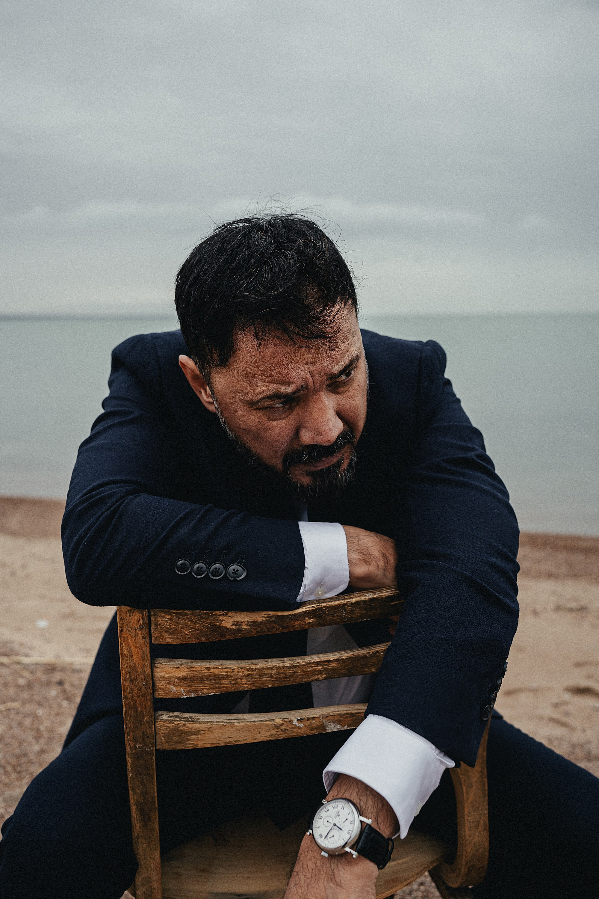 Мужчина в тёмном костюме сидит на деревянном стуле на песчаном берегу, задумчиво смотря вдаль