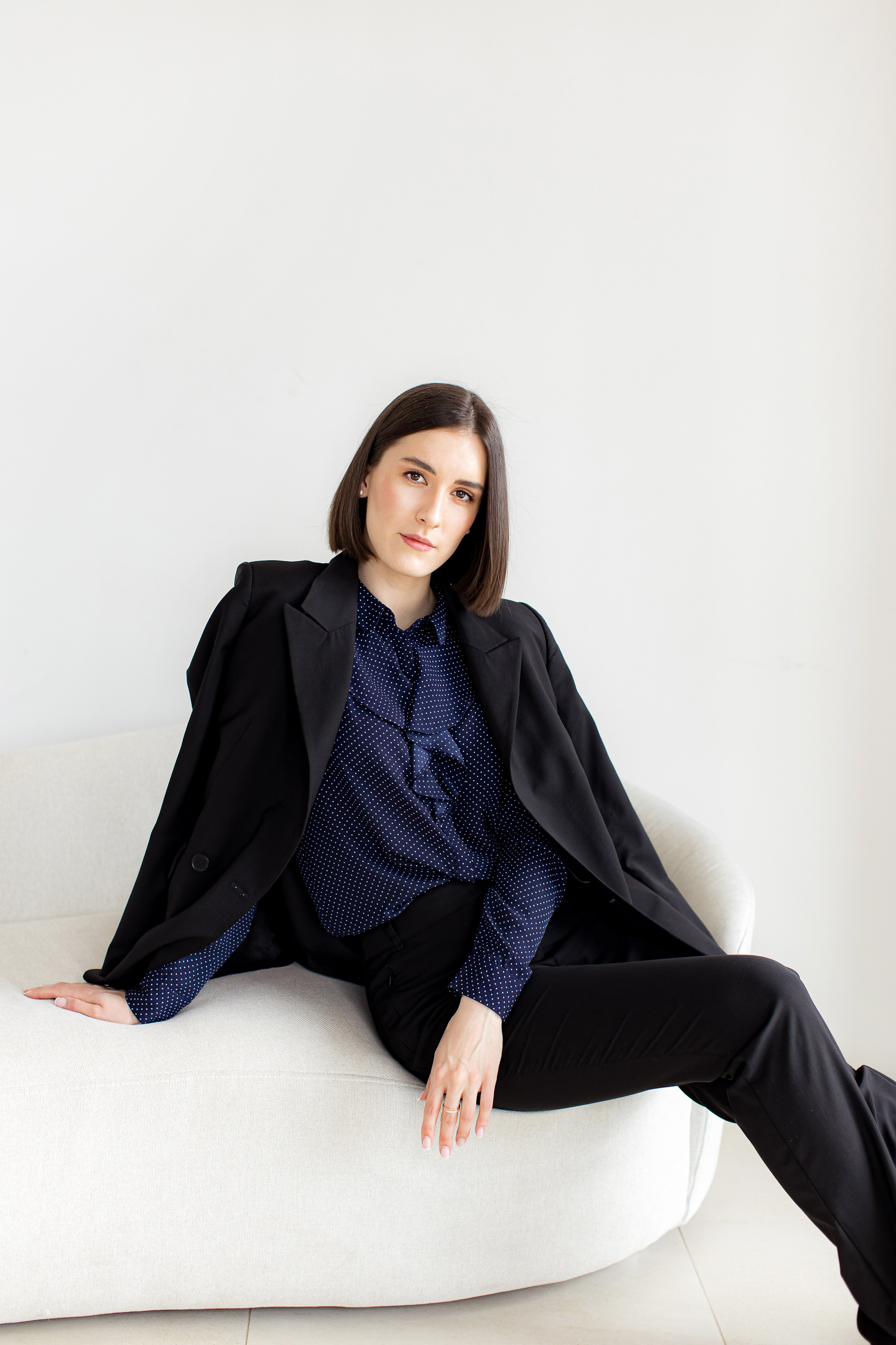 Девушка брюнетка в черном костюме и синей рубашке в горошек сидит на светлом диване