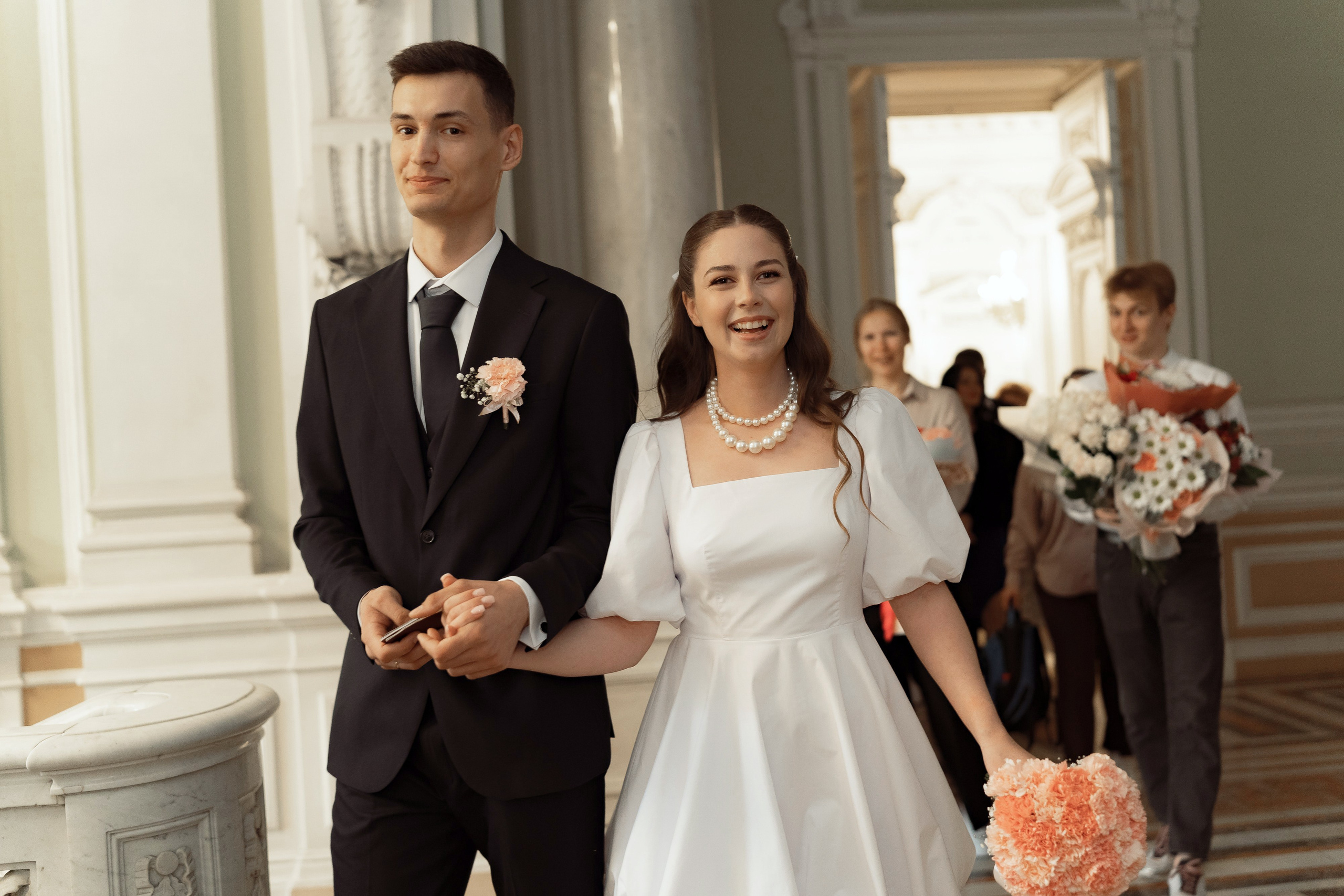 Жених и невеста с букетом улыбаются, уходя по коридору дворца