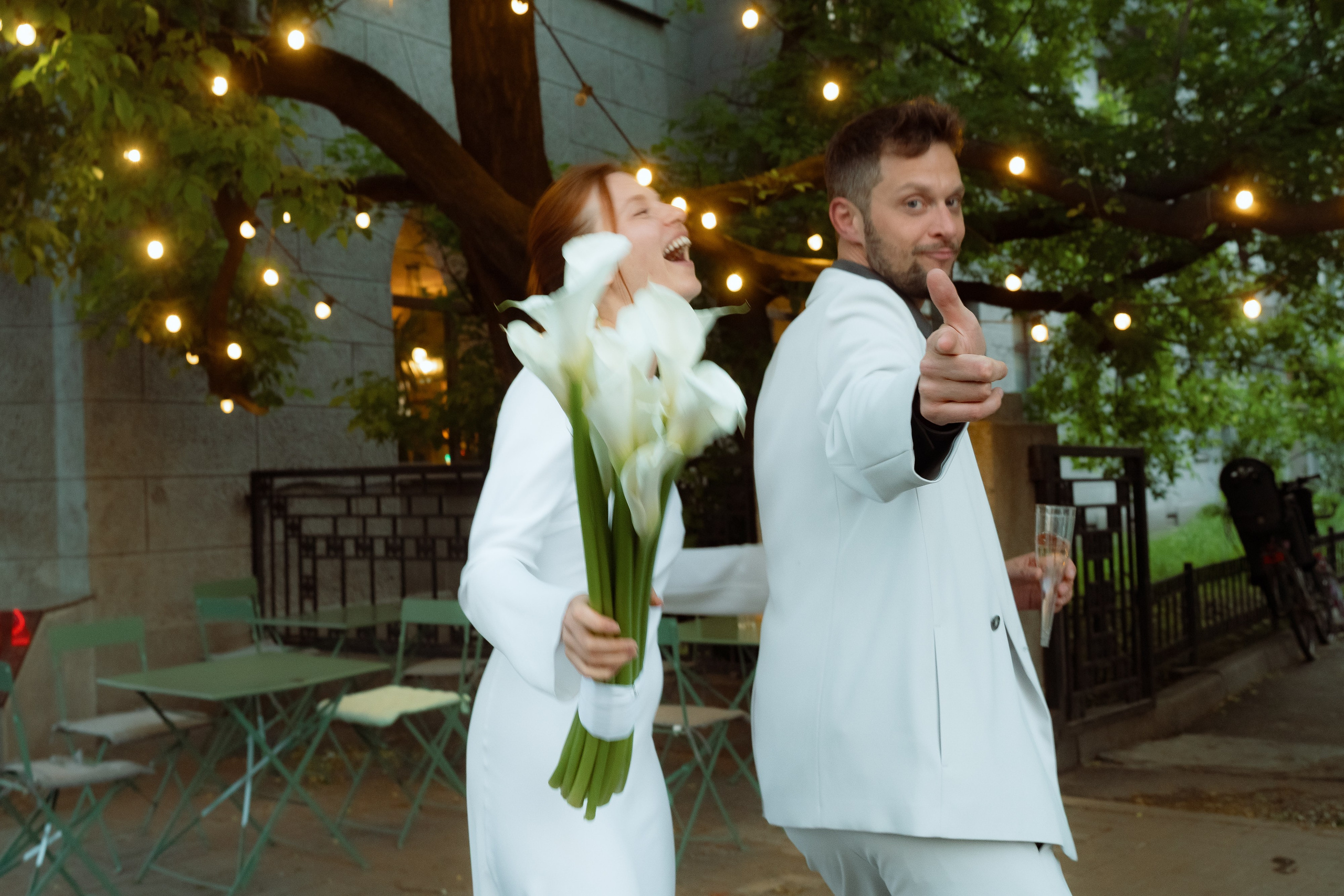 Мария с букетом калл смеётся, Евгений указывает вперёд под гирляндами огней