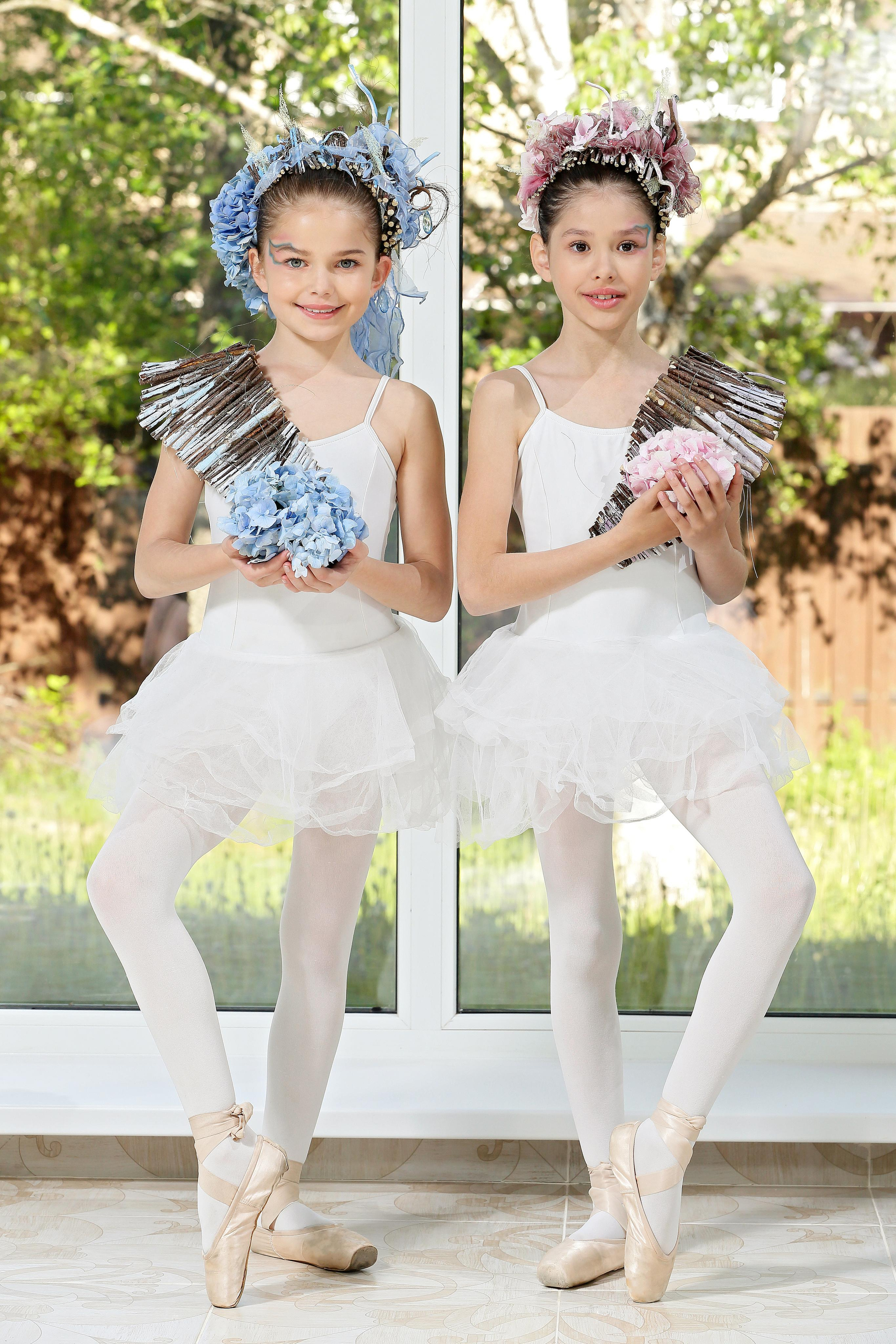 Две девочки-балерины в белых пачках с цветочными украшениями голубого и розового цвета на фоне окна.