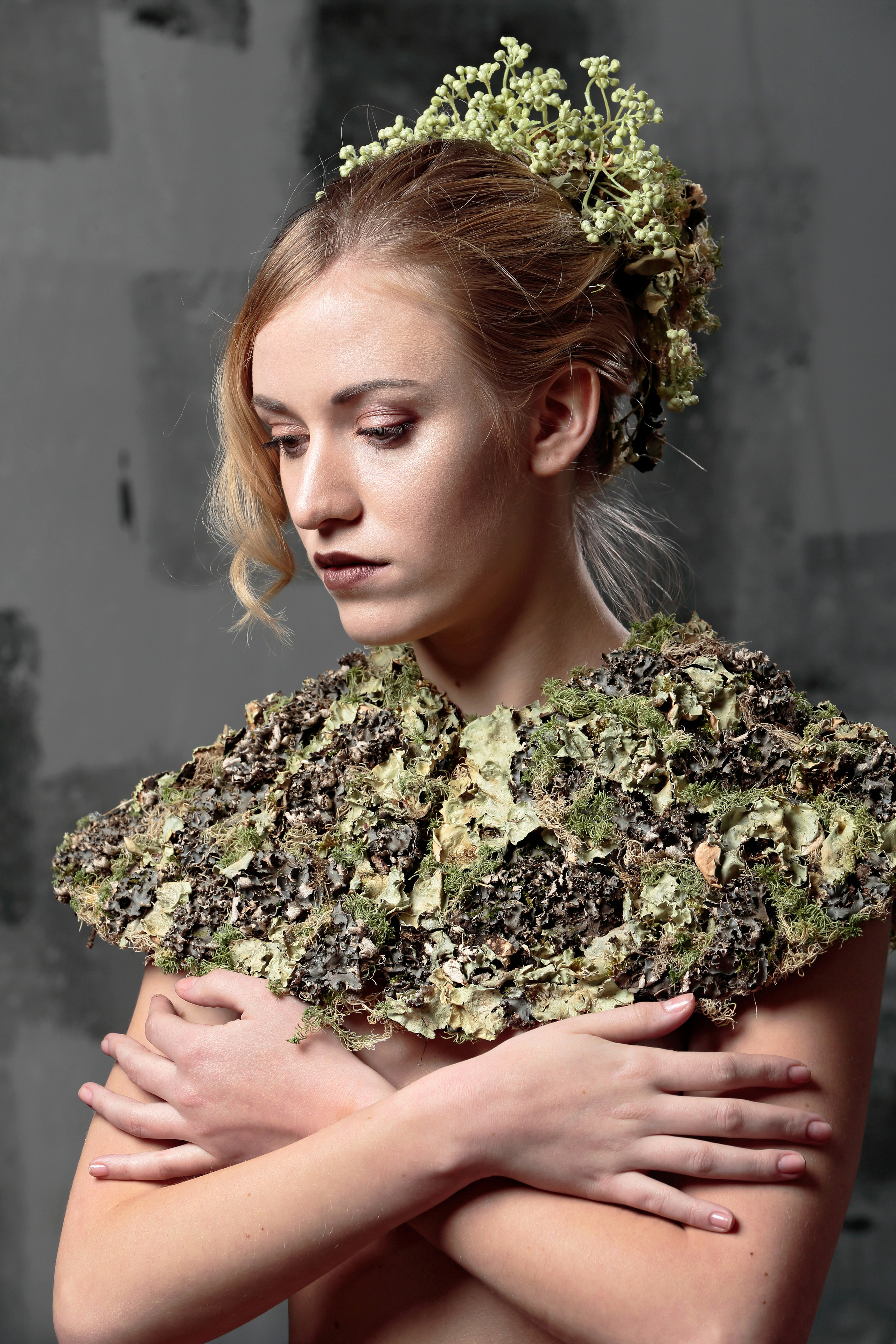 Поясной портрет молодой девушки в цветочном воротнике и цветочном украшении на голове на фоне серой стены.