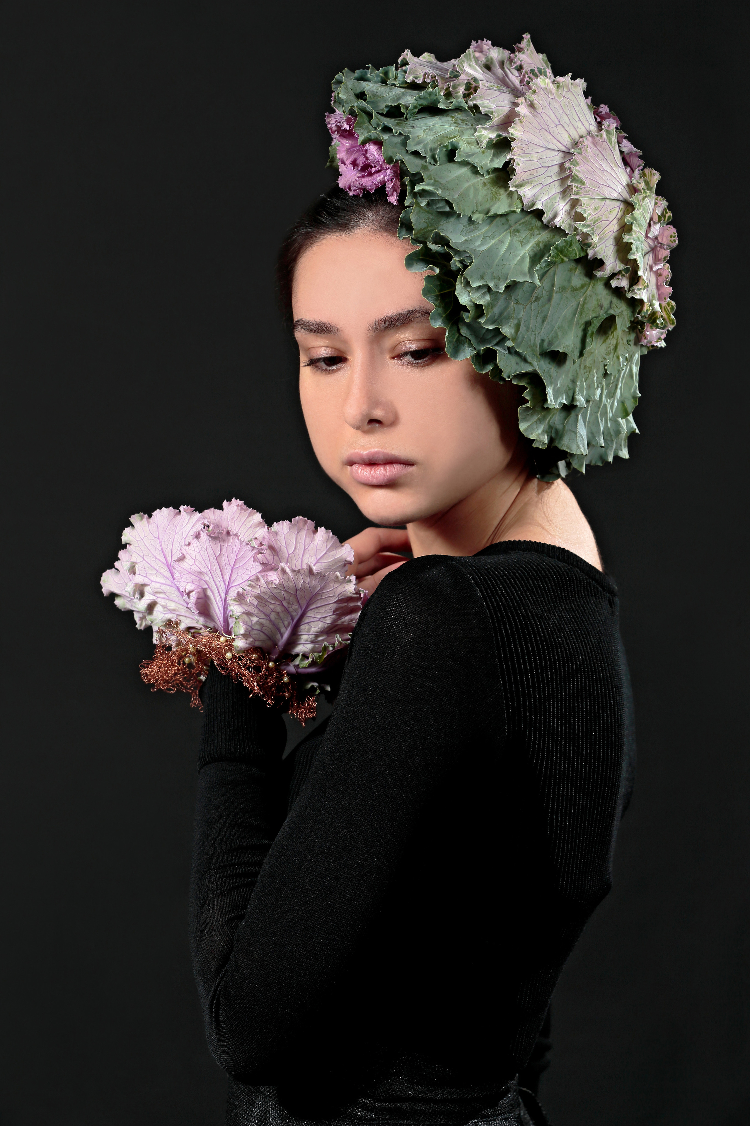 Девушка с головным убором и украшением на руке из листьев капусты на чёрном фоне.