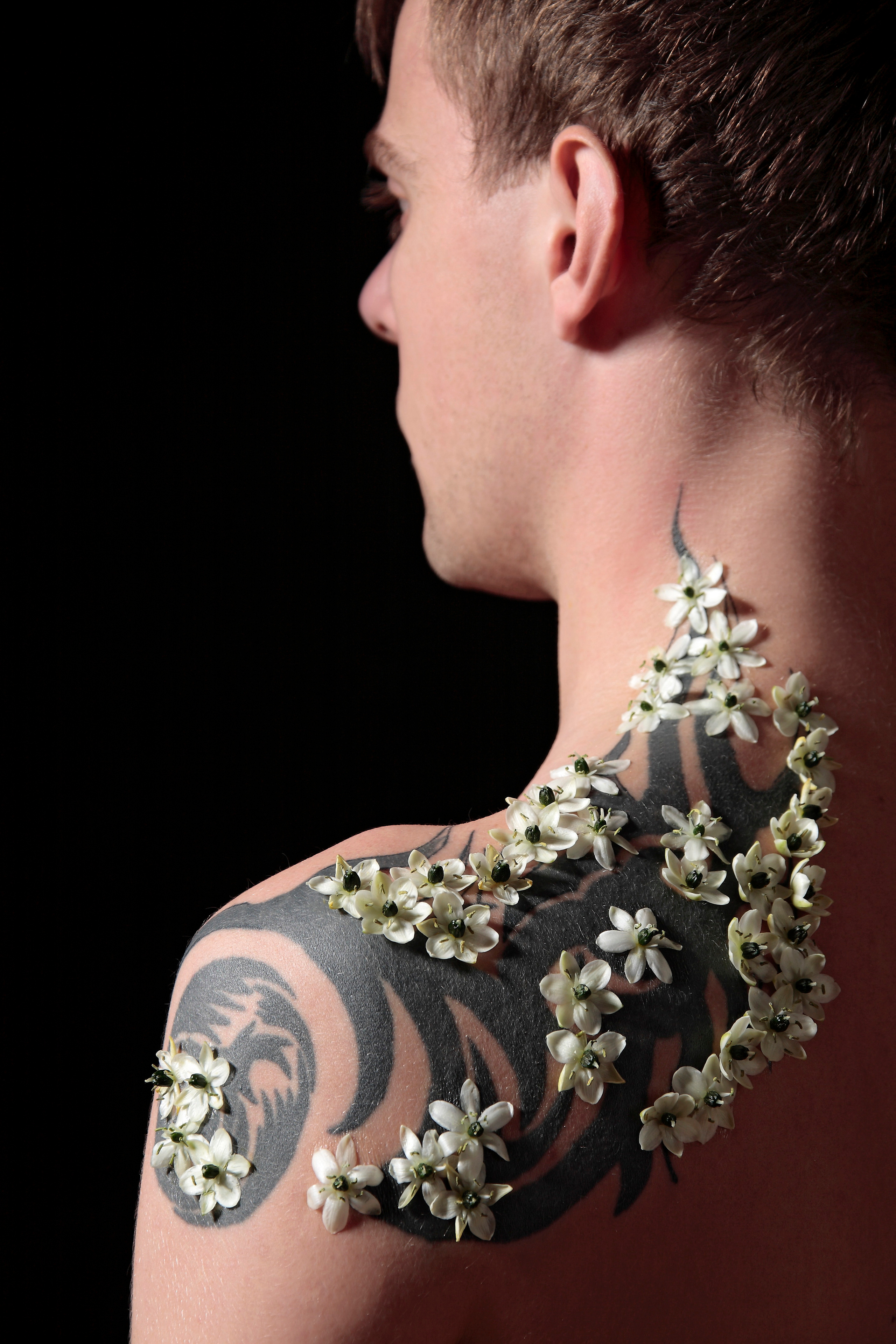 Молодой человек с татуировкой на плече, украшенной белыми цветами  на чёрном фоне.