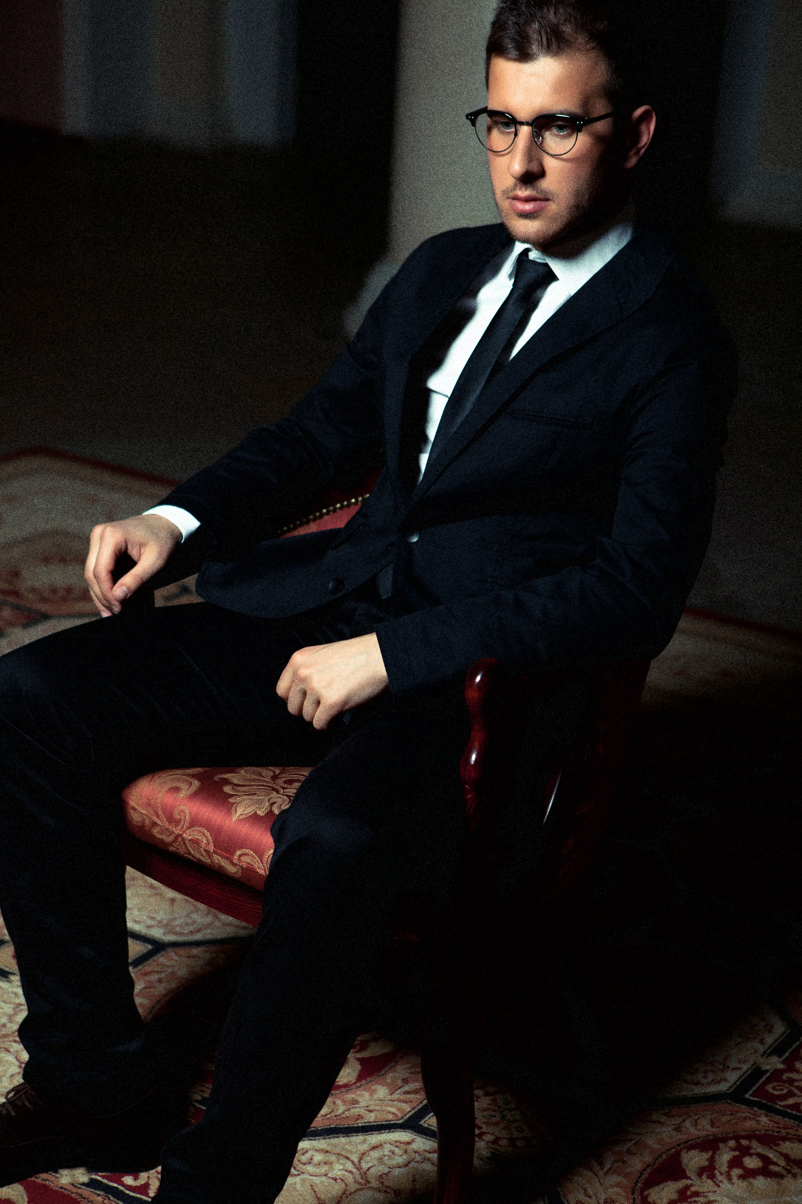 Мужчина в деловом костюме с черным галстуком и в очках сидит на стуле