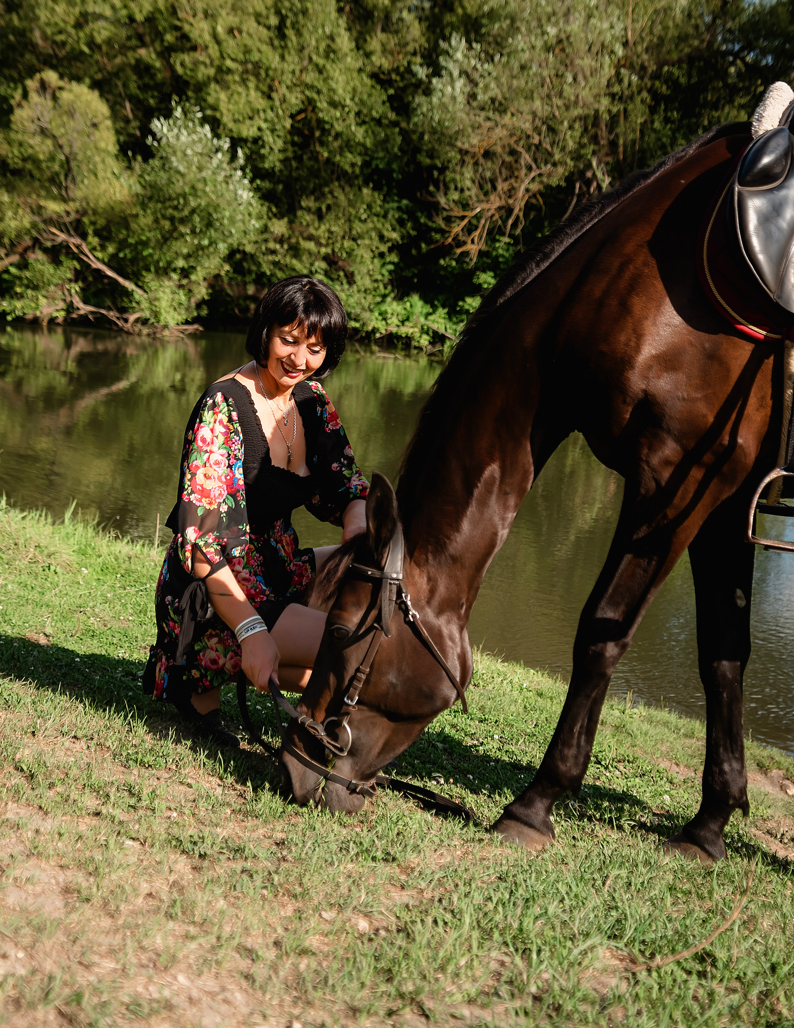 На фото женщина в бежевом платье стоит с лошадью на опушке леса.