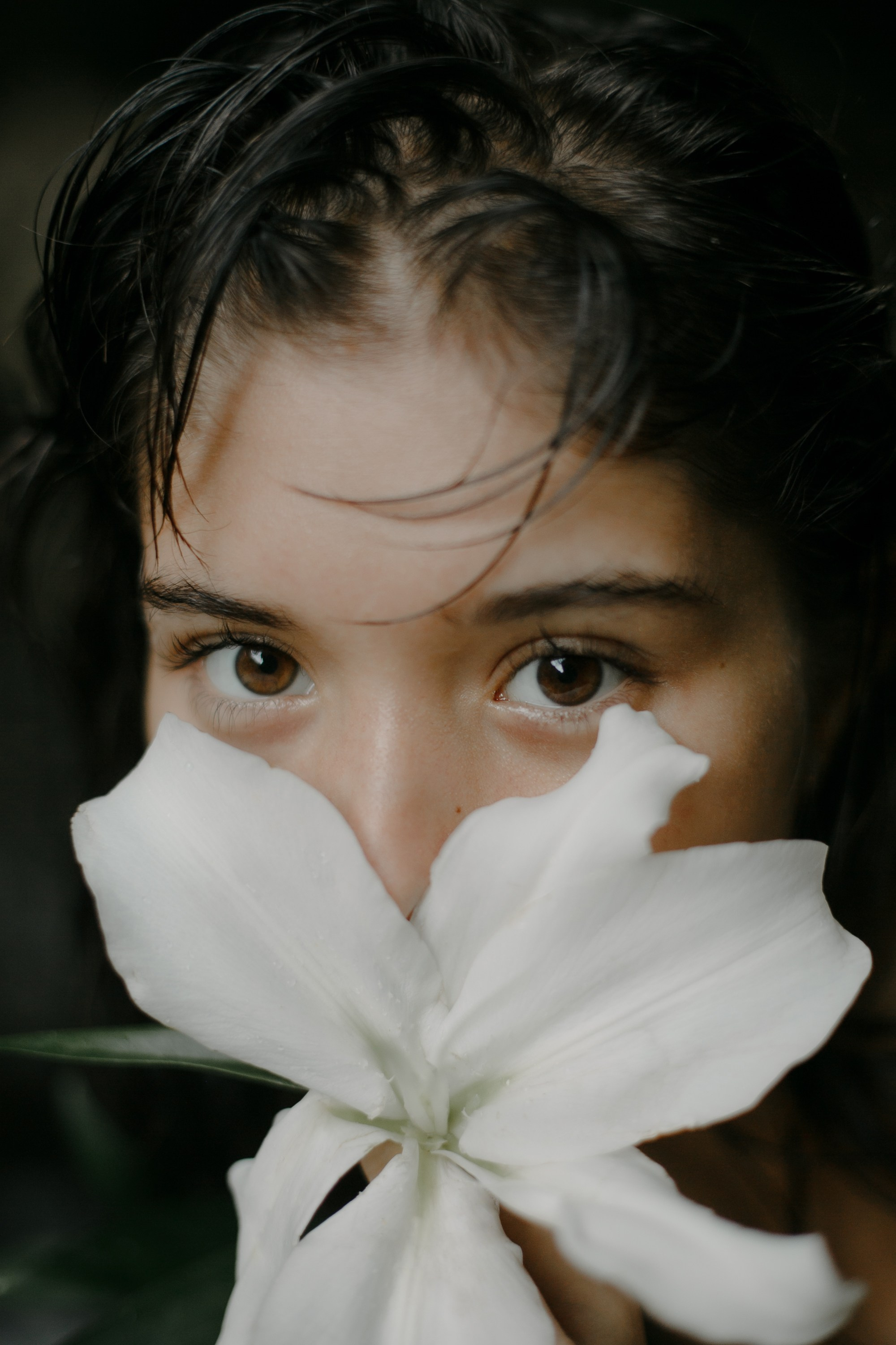 Крупный план девушки с влажными волосами, закрывающей лицо белым цветком. Естественный и загадочный образ с акцентом на глаза. Close-up of a girl with wet hair, her face partially covered by a white flower. A natural and mysterious look with a focus on the eyes.