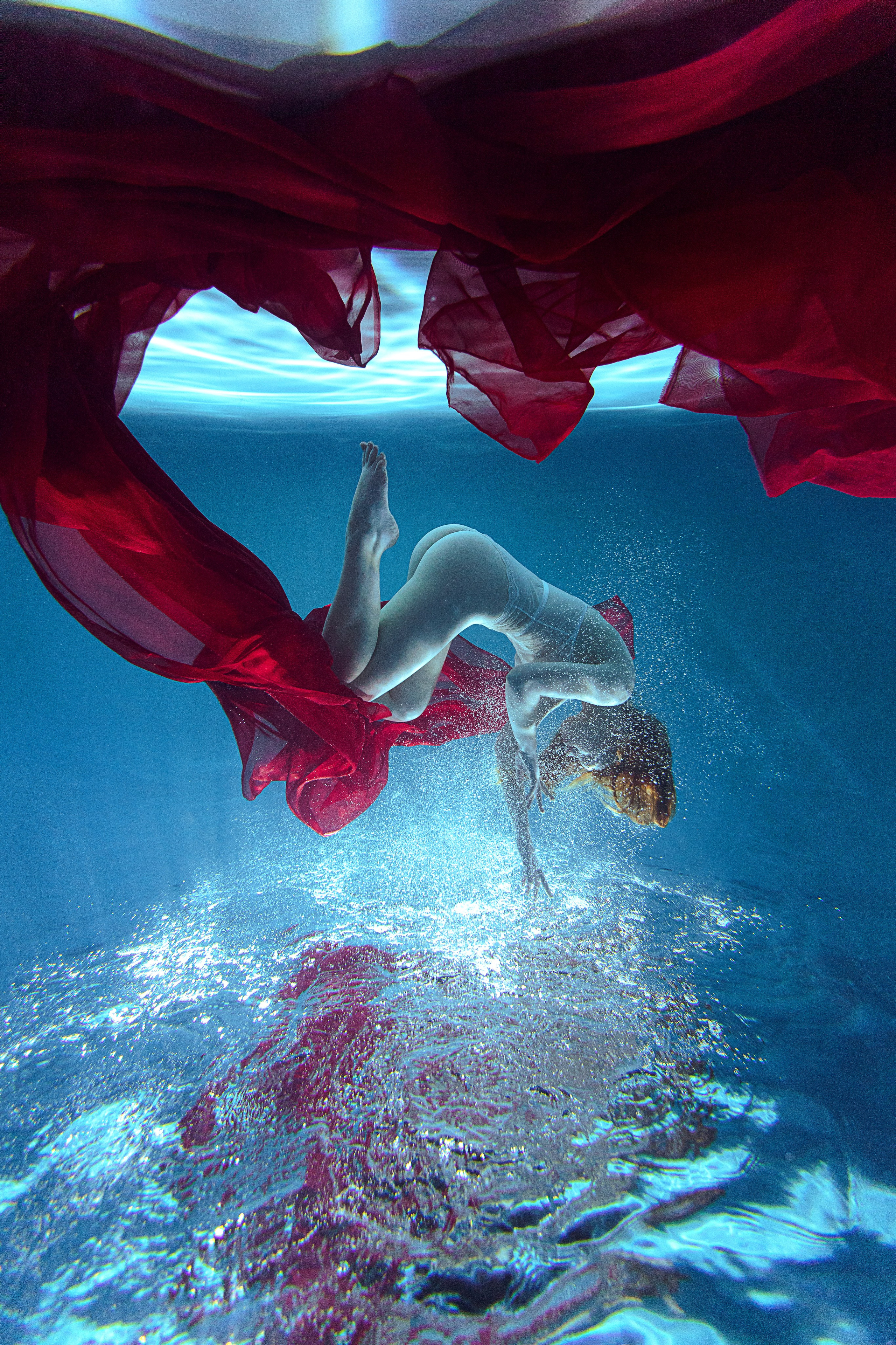 Подводная съёмка: модель плавно движется под водой, её тело окутано струящейся красной тканью. Свет мягко преломляется в воде, создавая ощущение невесомости и художественной грации. Атмосфера кадра — мистическая и эстетичная, подчёркивающая плавность движения и игру цвета