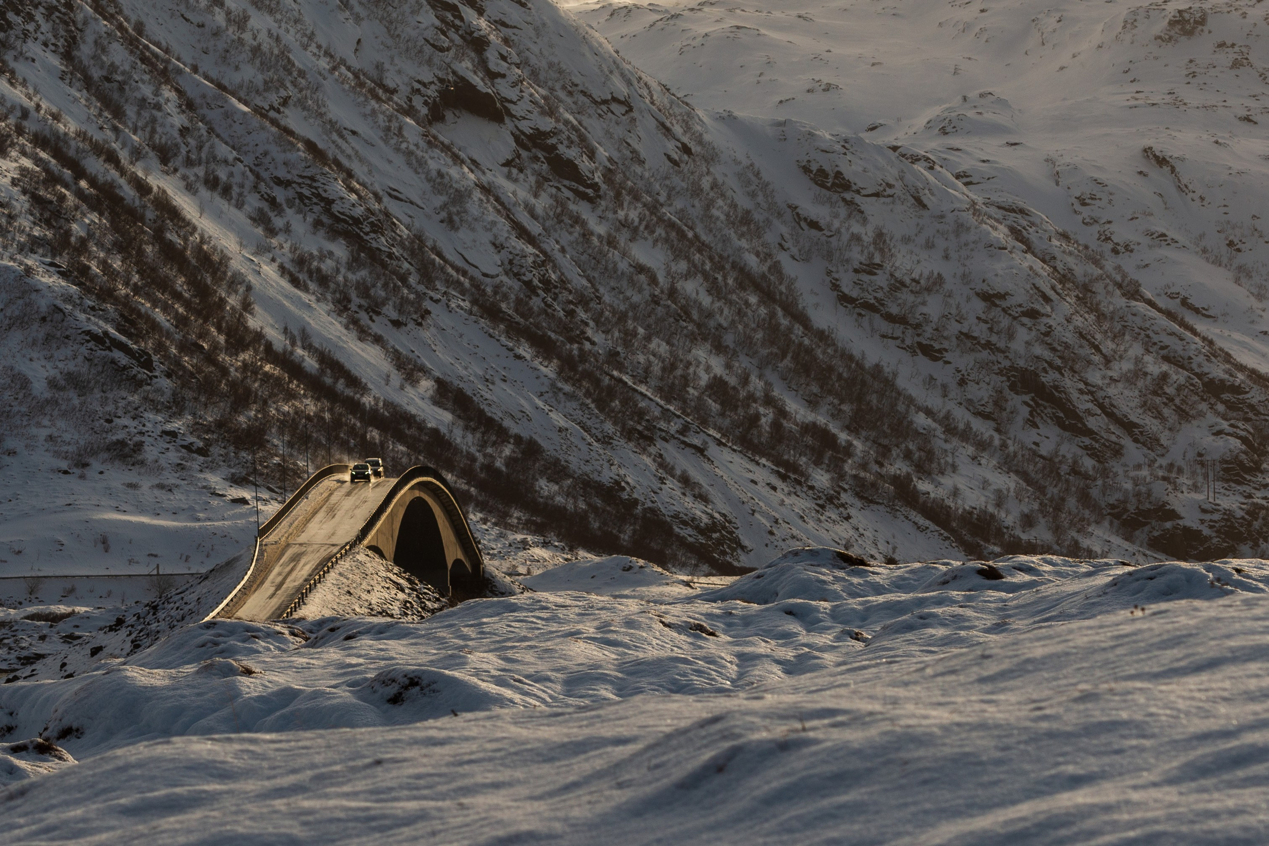 Зимний пейзаж Лофотенских островов в Норвегии с изображением Сторсезандетского моста (Storseisundet brua) — знаменитого консольного моста, который является частью живописной Атлантической дороги (Atlanterhavsveien). Мост изгибается над заснеженным ландшафтом, соединяя острова, на фоне величественных снежных гор.