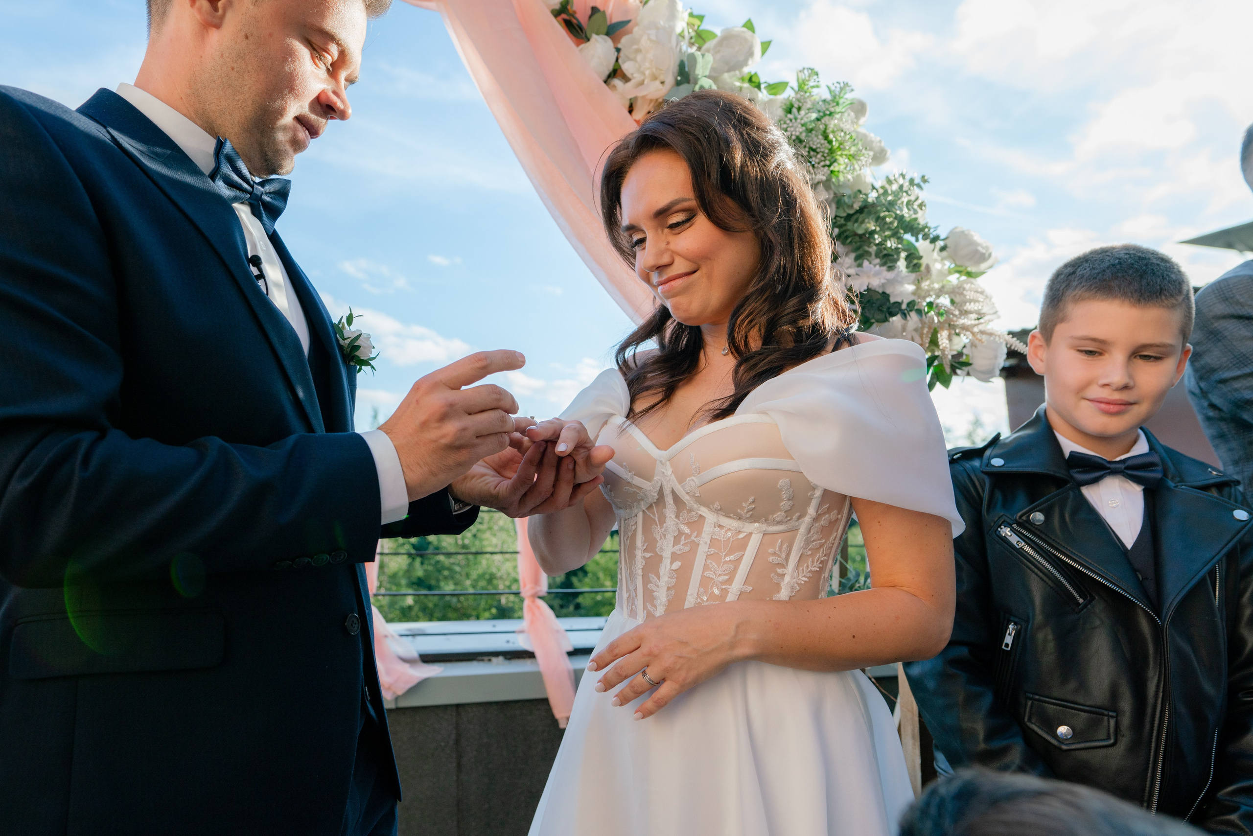 Жених в синем костюме надевает кольцо на невесту в белом платье на выездной регистрации на шоу Четыре свадьбы