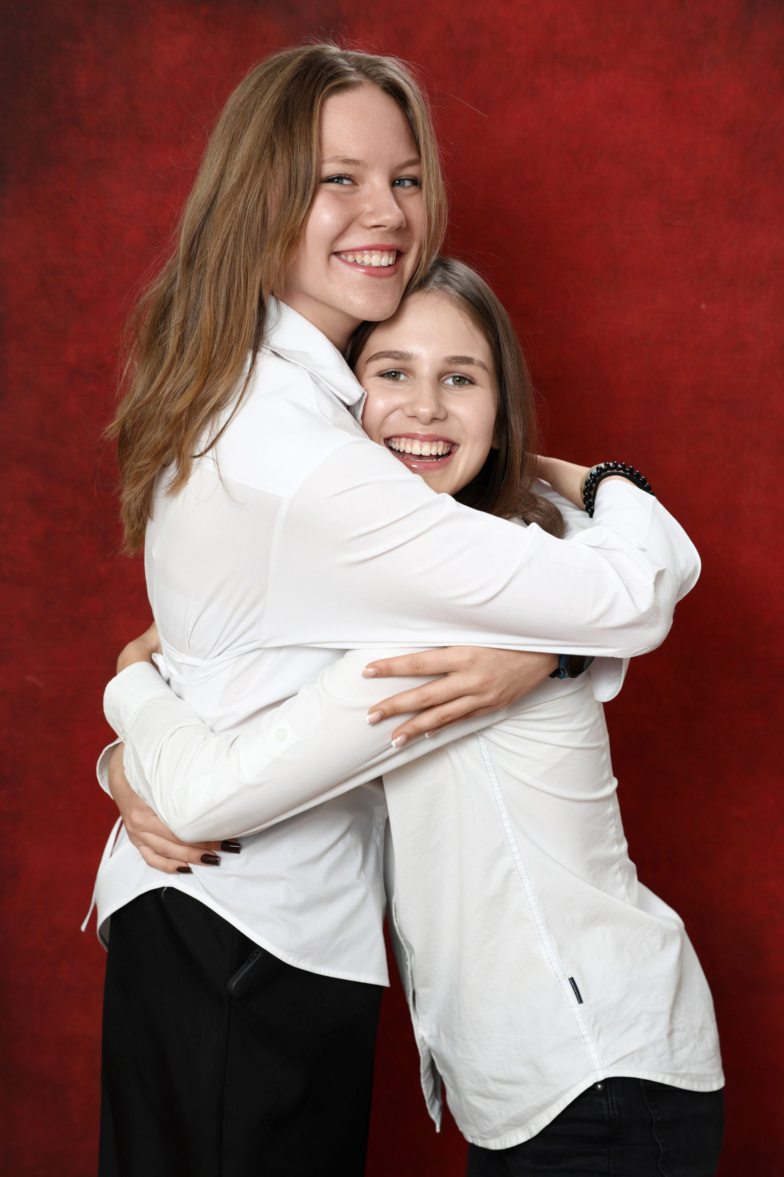 Две девушки в светлых рубашках обнимаются в парном кадре на красном фоне.