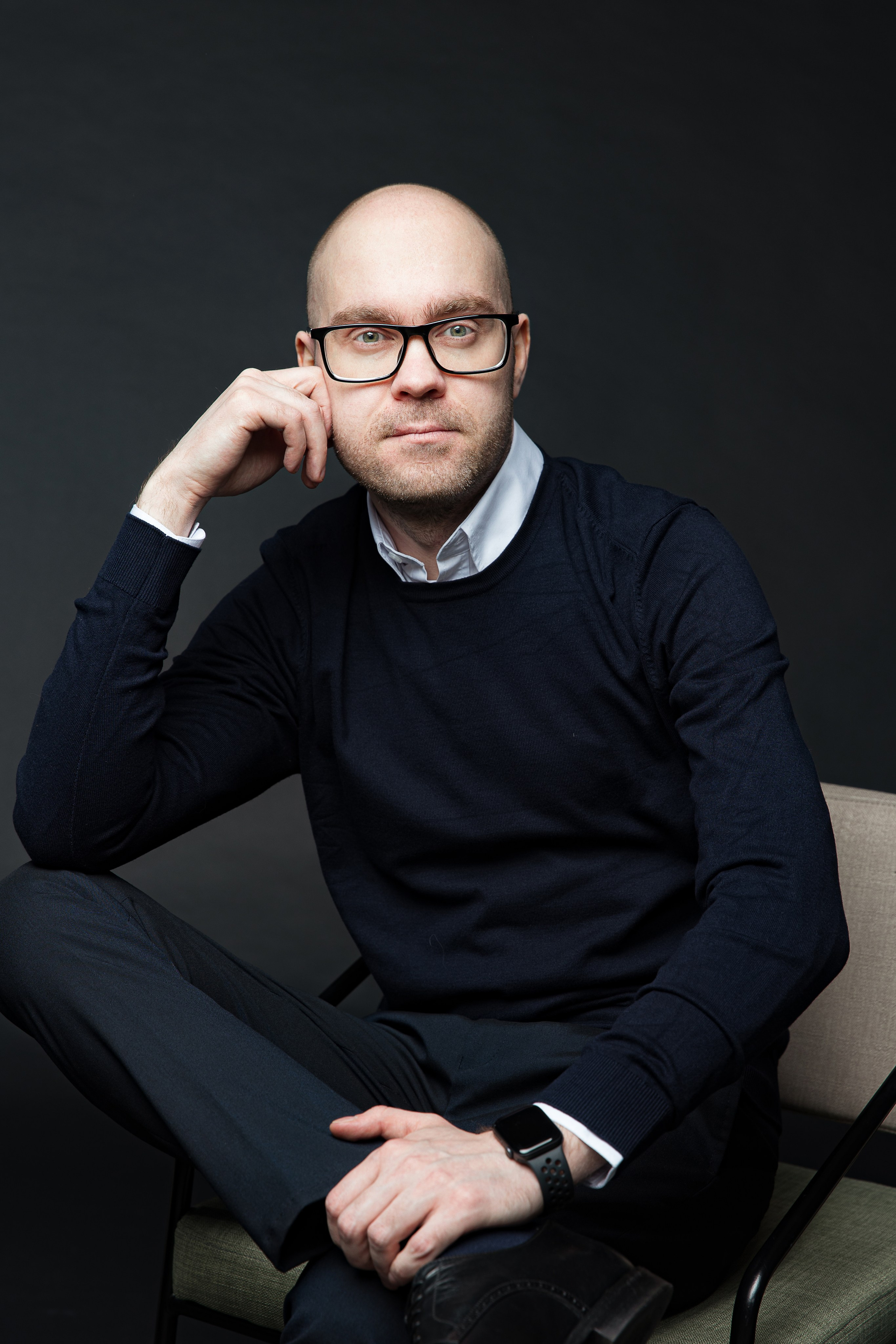 Лысый мужчина в джемпере и рубашке сидит на стуле на темном фоне