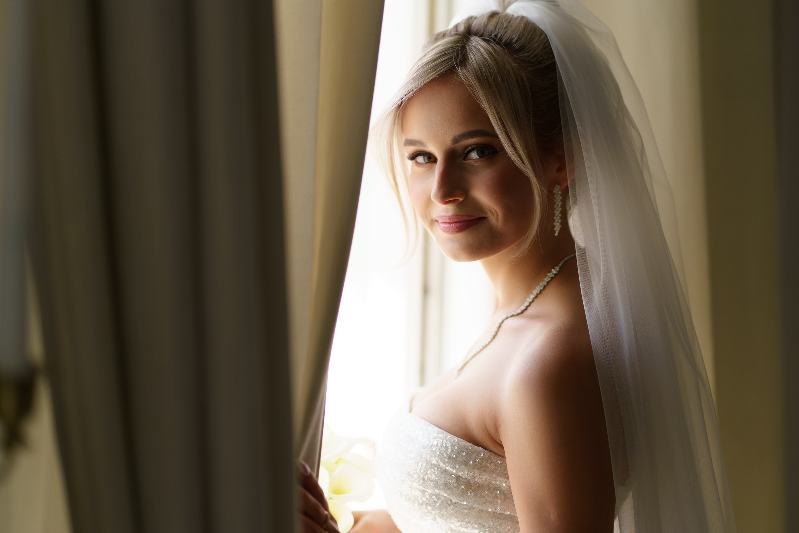 Невеста в белом платье с блестящими деталями смотрит через плечо. На ней длинная фата, длинные серьги и ожерелье. Атмосфера снимка очень романтична, свет мягко ложится на её лицо | фотосессия для молодоженов Екатеринбург