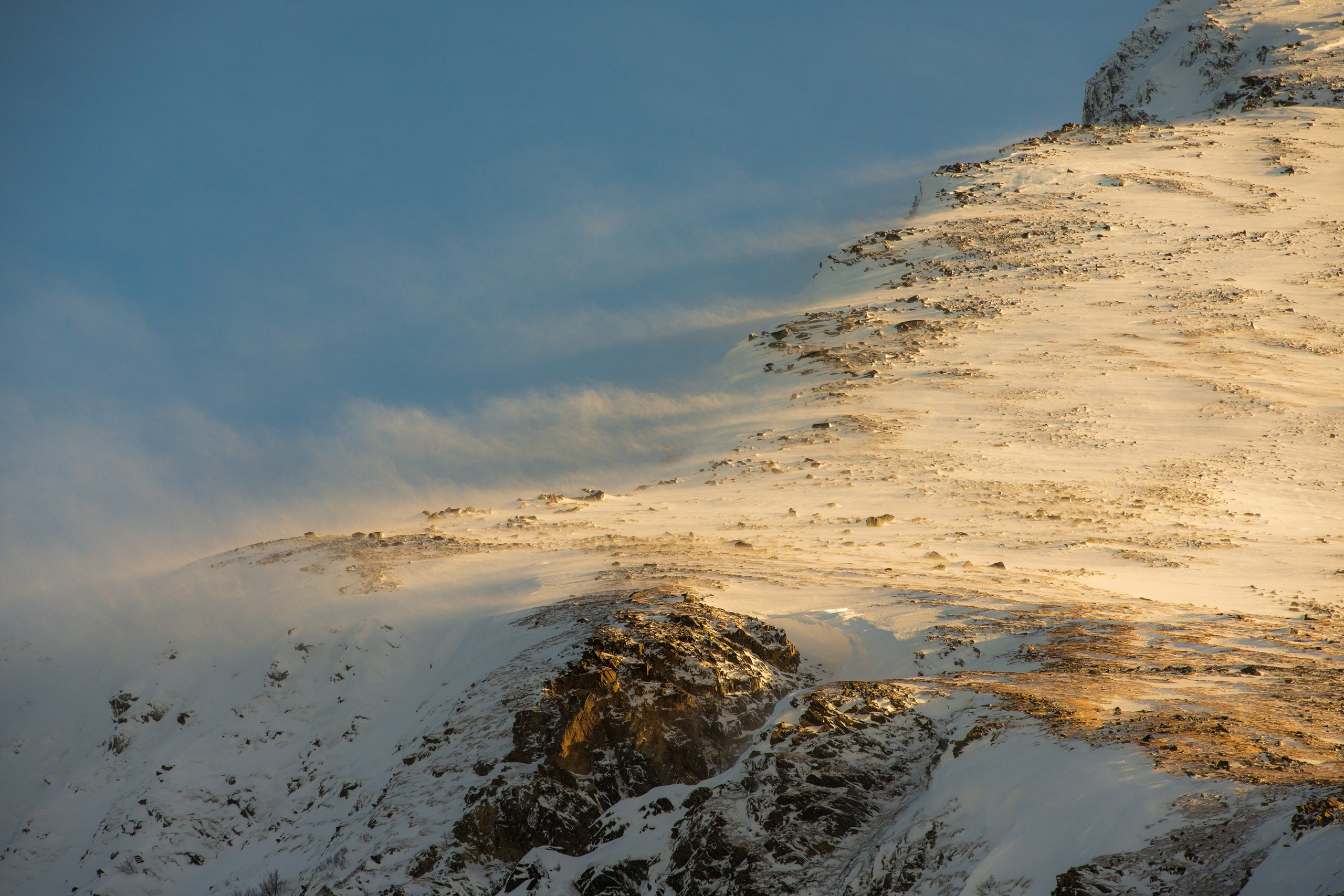 Заснеженный горный склон, освещенный низким золотистым солнцем, с сильным ветром, поднимающим снег.