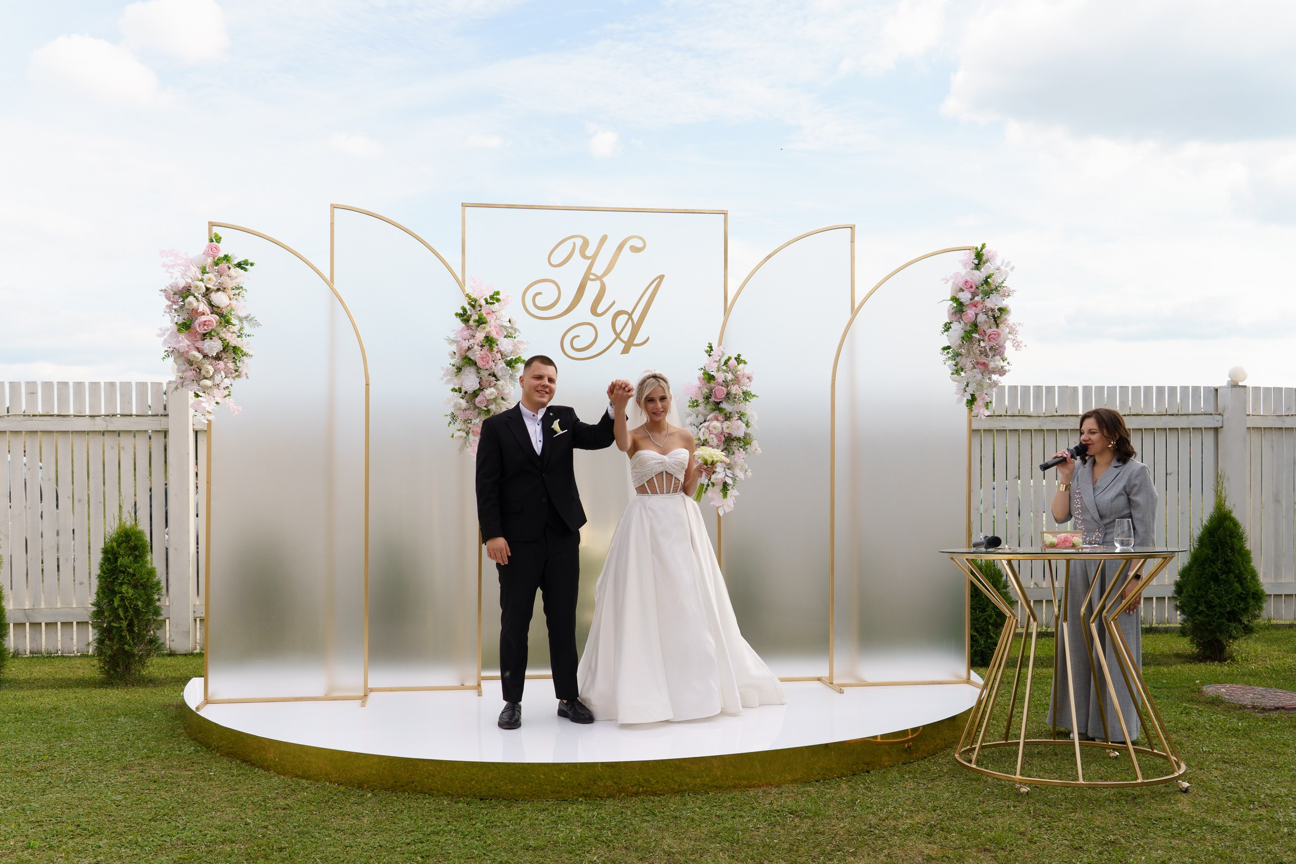 Изящная свадебная церемония: молодожёны стоят на круглой сцене с золотыми элементами, окружённой пышными букетами розовых и белых цветов. Ведущая с микрофоном готова продолжить праздник. Атмосфера наполнена радостью и любовью| свадебный фотограф для выездной регистрации Екатеринбург