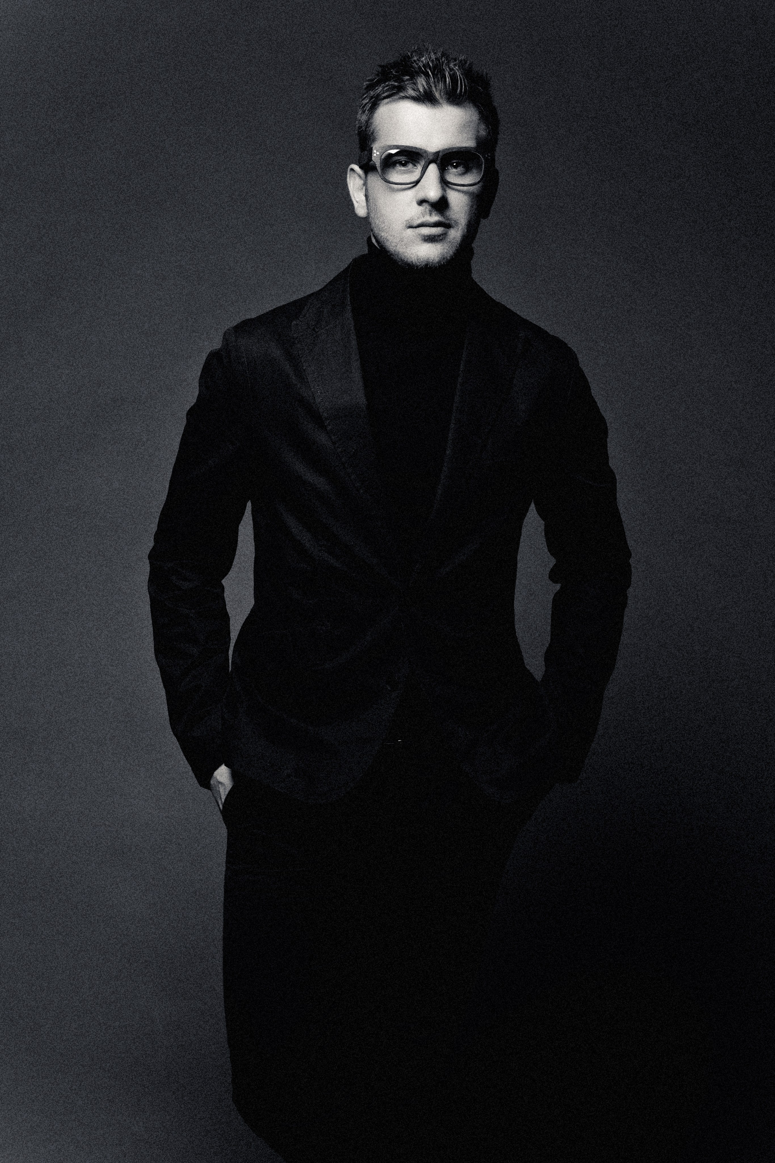 Стильный мужчина в костюме с водолазкой и в очках стоит на черном фоне