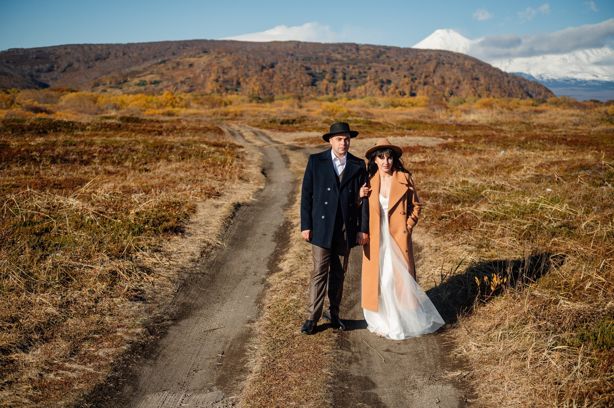 Пара в стильных пальто и шляпах стоит на просёлочной дороге. Вокруг — осенние поля, на фоне горы и снежная вершина. Атмосфера романтичная, немного дикая.
