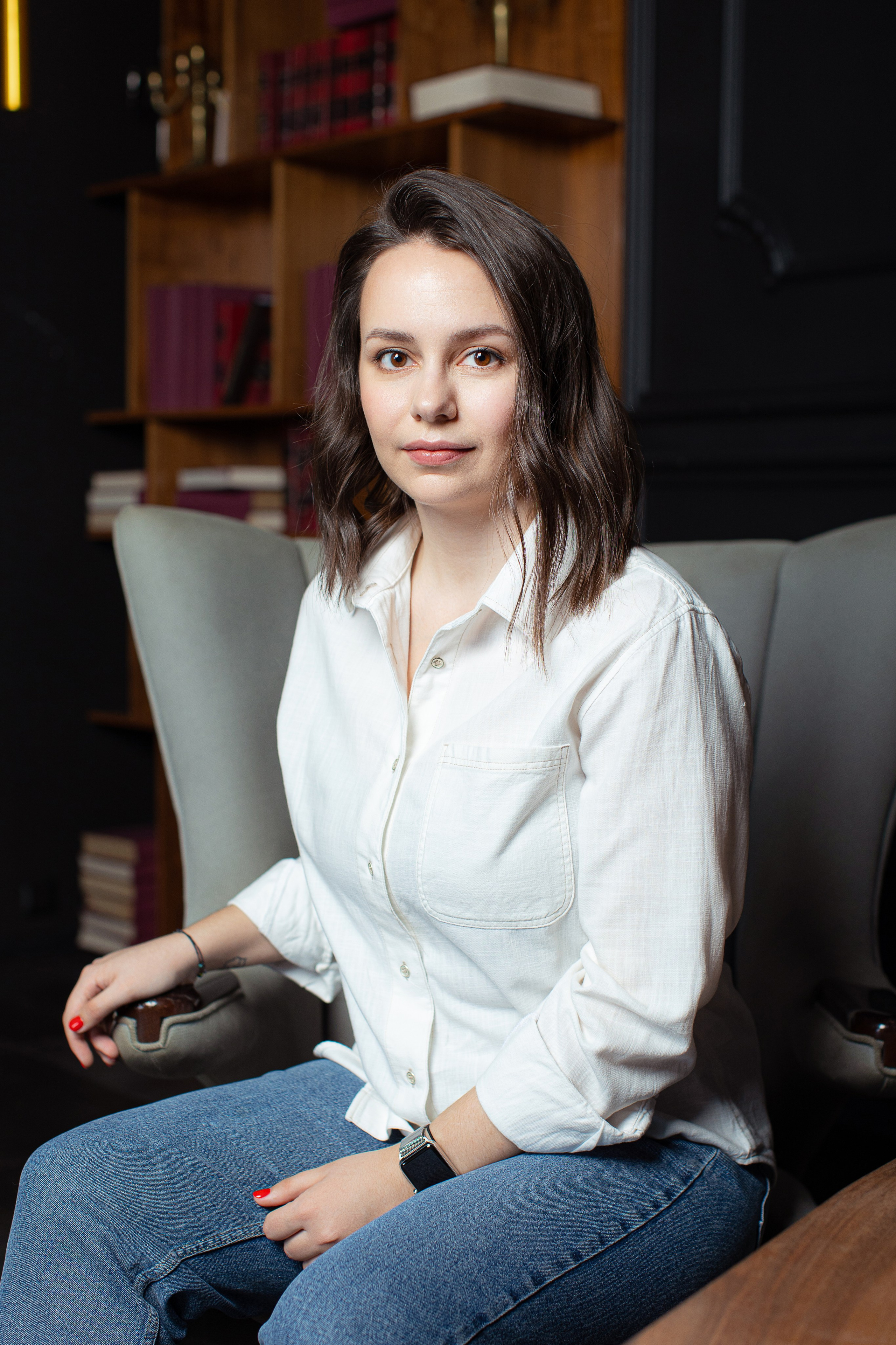 Девушка в белой рубашке и синих джинсах сидит в сером кресле в интерьере