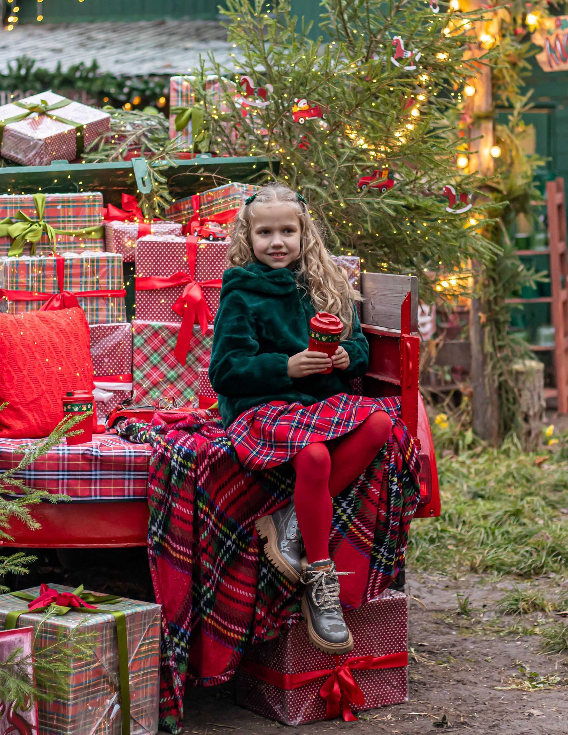 Девочка в зелёной шубке на фоне новогодней фотозоны с ёлками и огоньками, фотограф Карасёва Елена