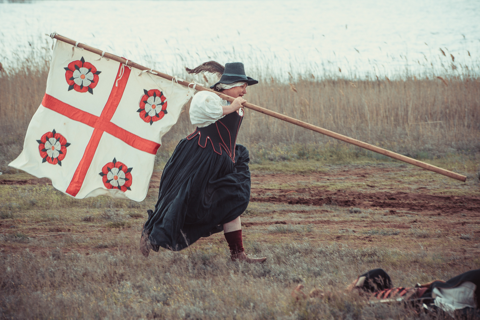 Женщина в старинном костюме бежит по полю со знаменем на плече