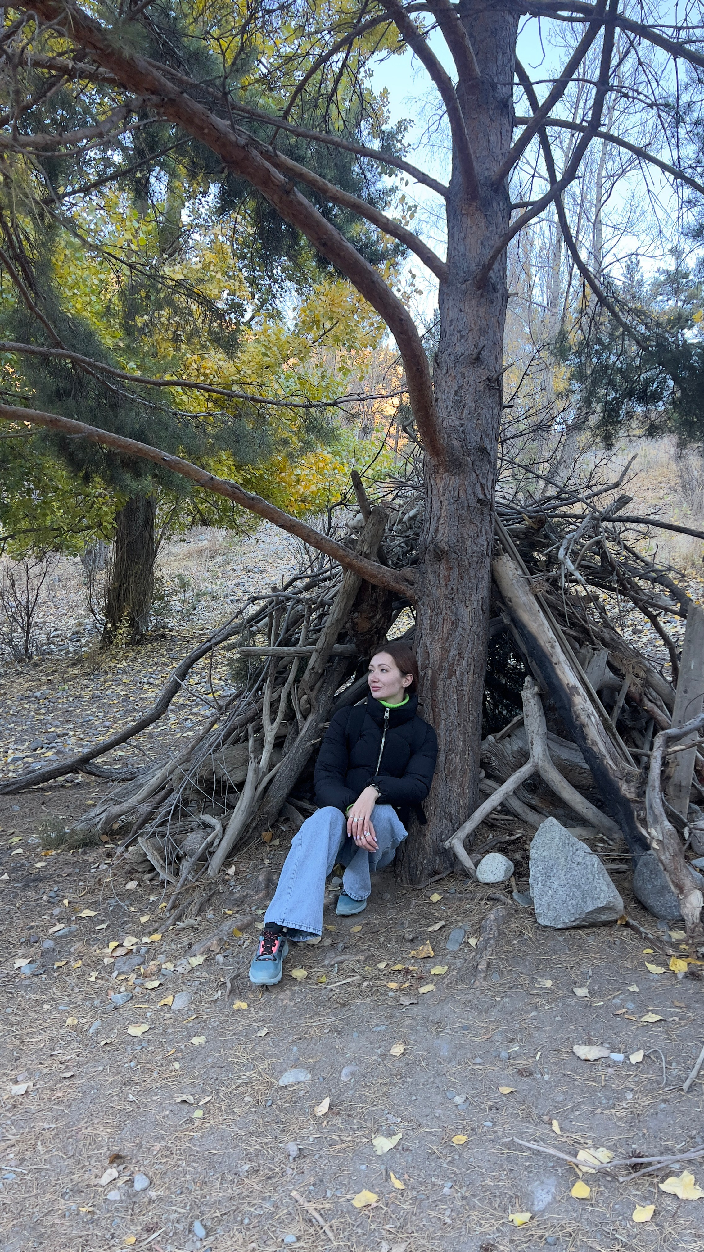 Молодая женщина в черной куртке и джинсах сидит у основания дерева на фоне импровизированного шалашика из веток на берегу озера Иссык. Вокруг осенний лес с пожелтевшими листьями и солнечным светом, пробивающимся сквозь ветви. Обстановка выглядит спокойной и умиротворяющей.
