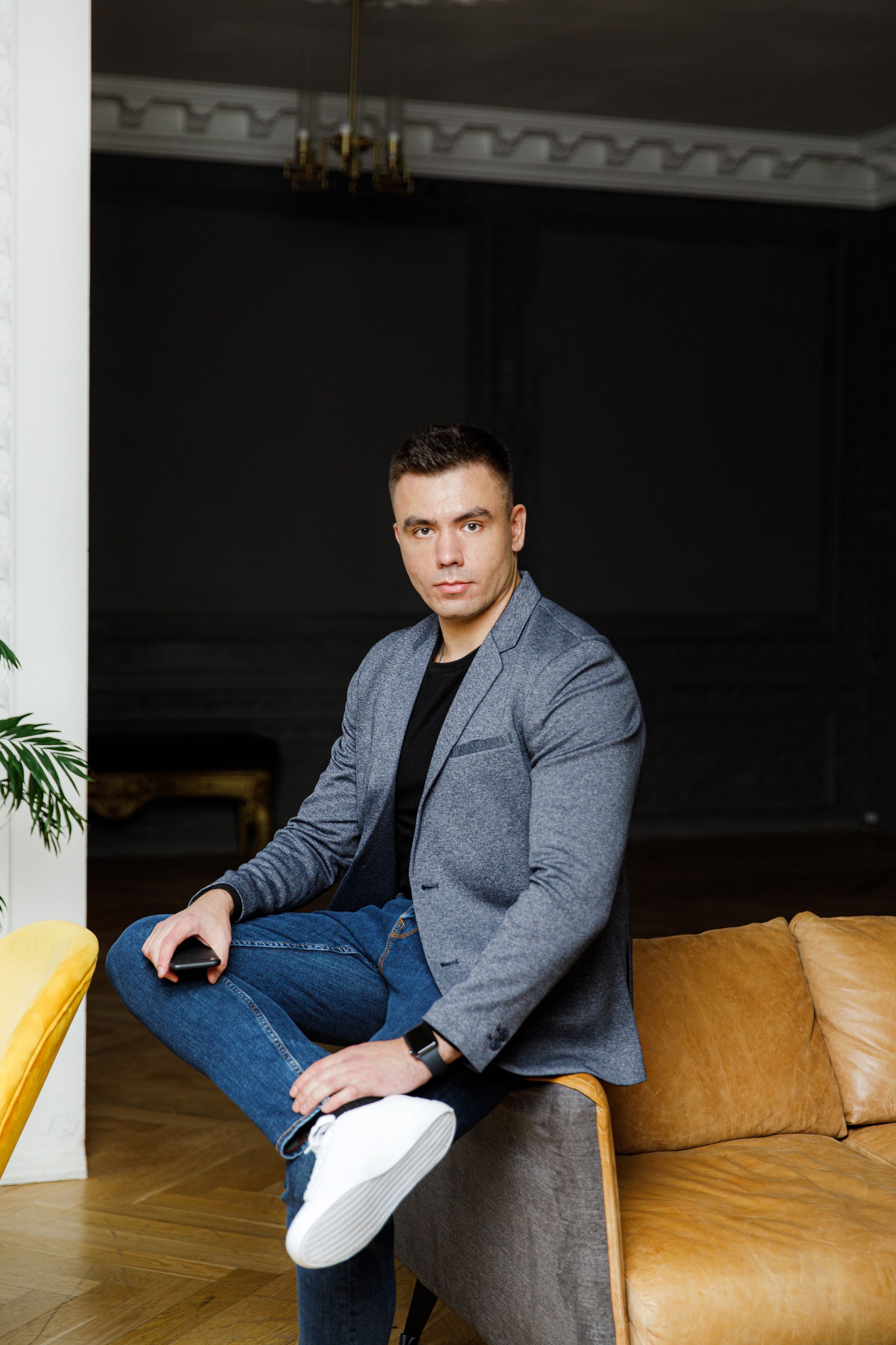 Мужчина в синих джинсах и сером пиджаке сидит на краю дивана