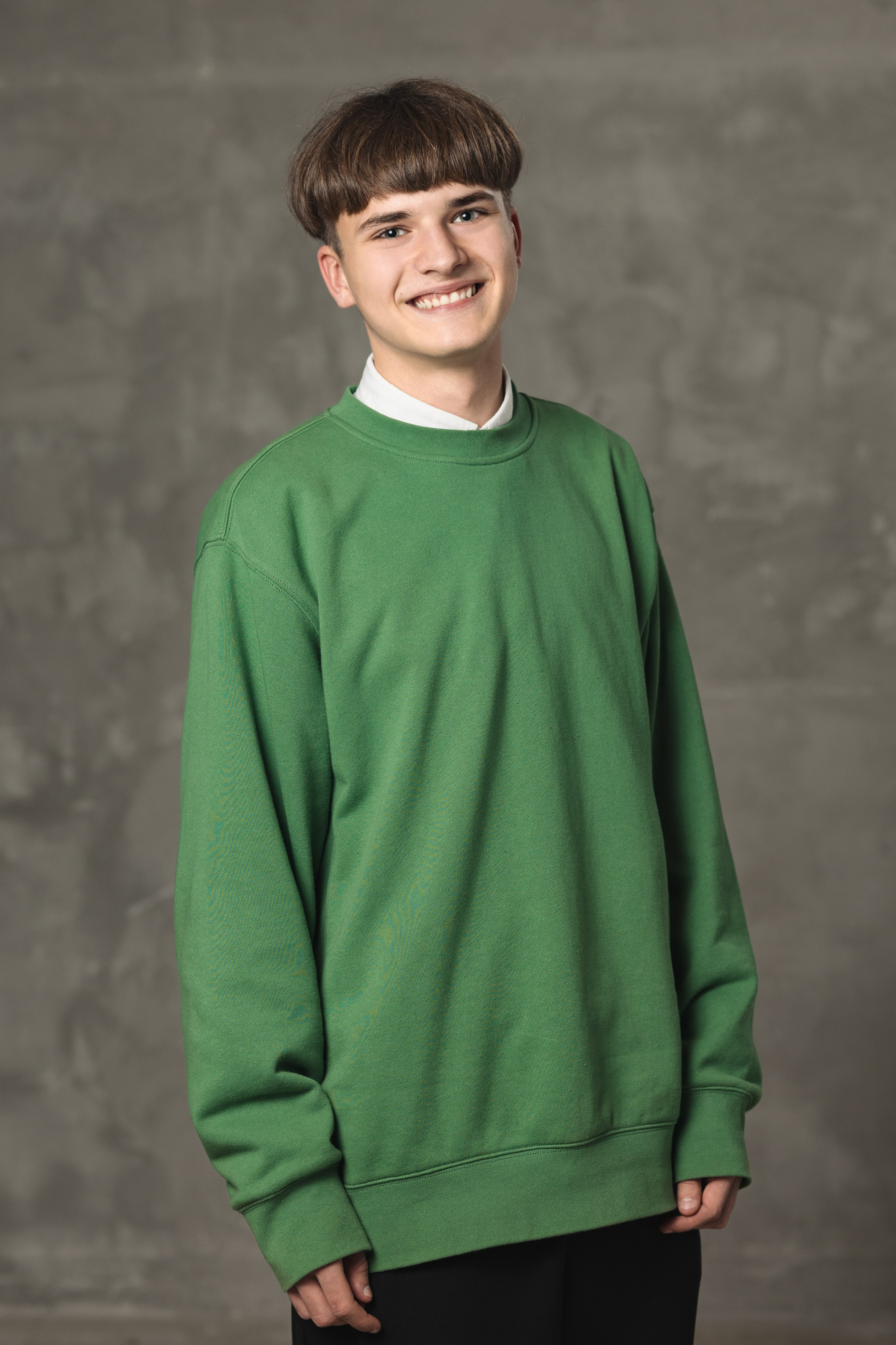 Ученик в зелёном свитере позирует на нейтральном сером фоне.