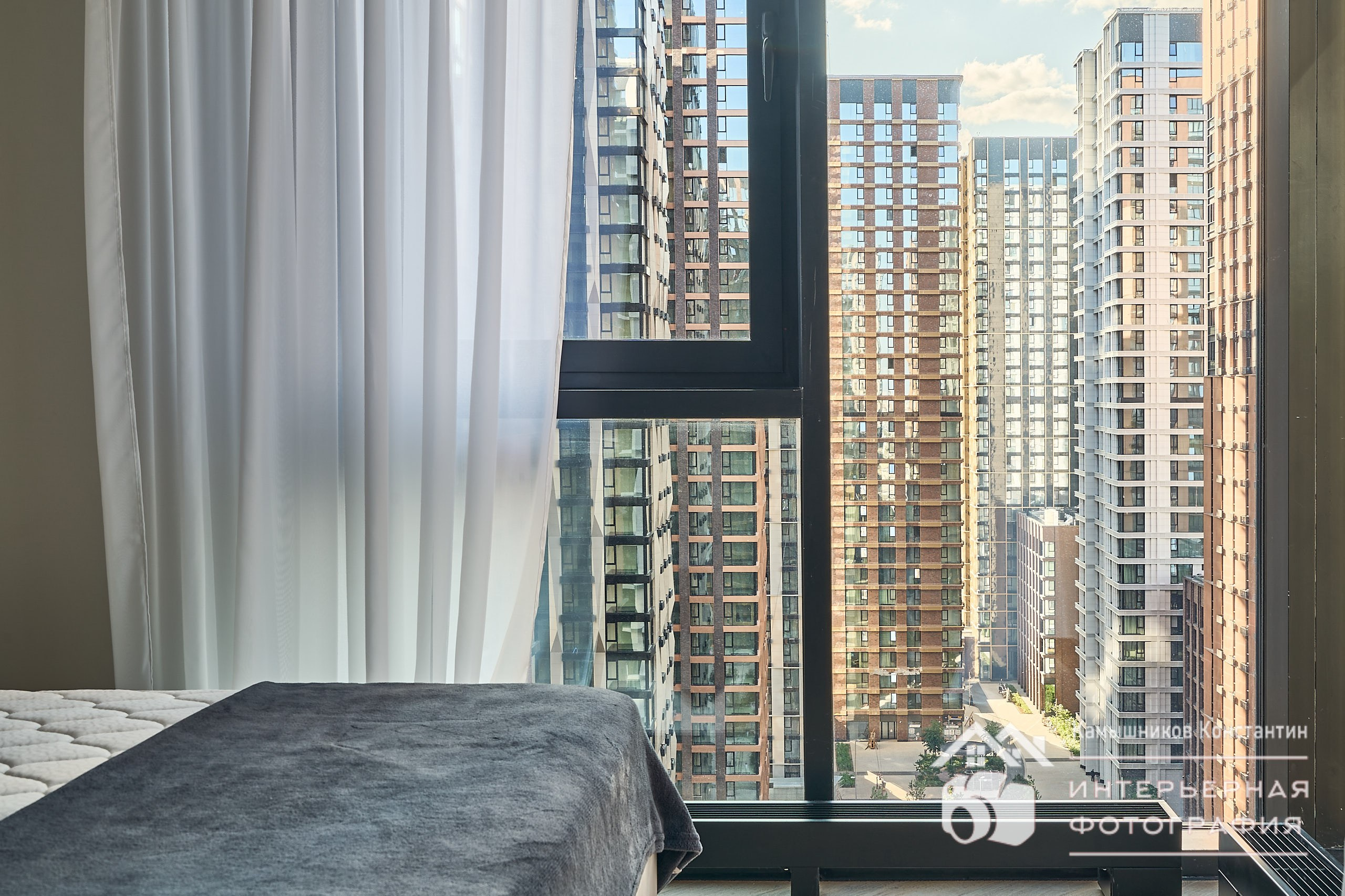 Интерьерная съемка апартаментов в ЖК Метрополия, небольшая уютная теплая спальня с огромным панорамным окном во всю стену, в кадре часть кровати и шикарный вид из окна