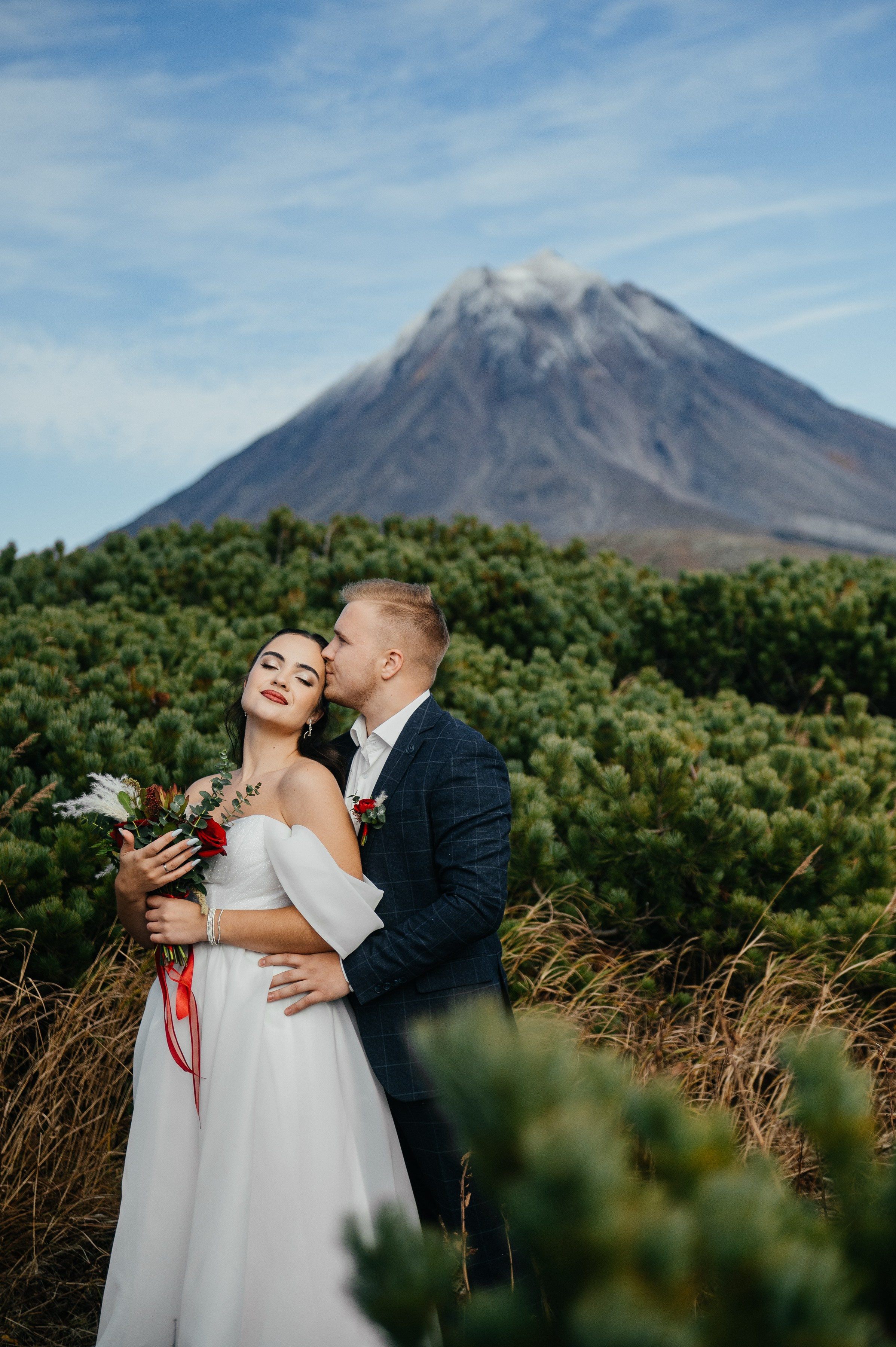 Жених в костюме обнимает невесту в белом платье, она держит букет. На фоне — вулкан и хвойные кусты. Атмосфера романтичная.
