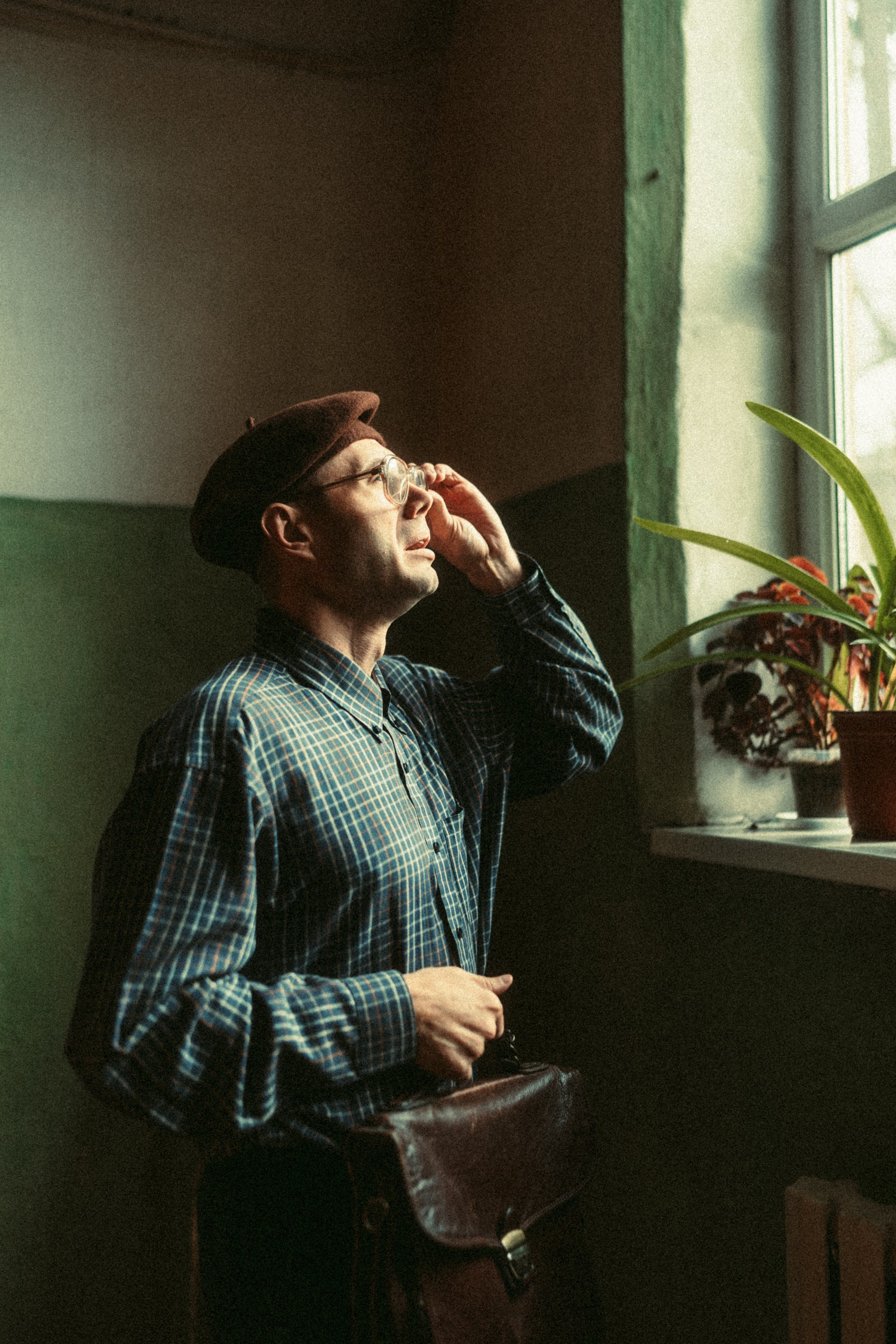 Мужчина в берете и клетчатой рубашке смотрит в окно на лестничной клетке в старом здании, освещённый дневным светом.