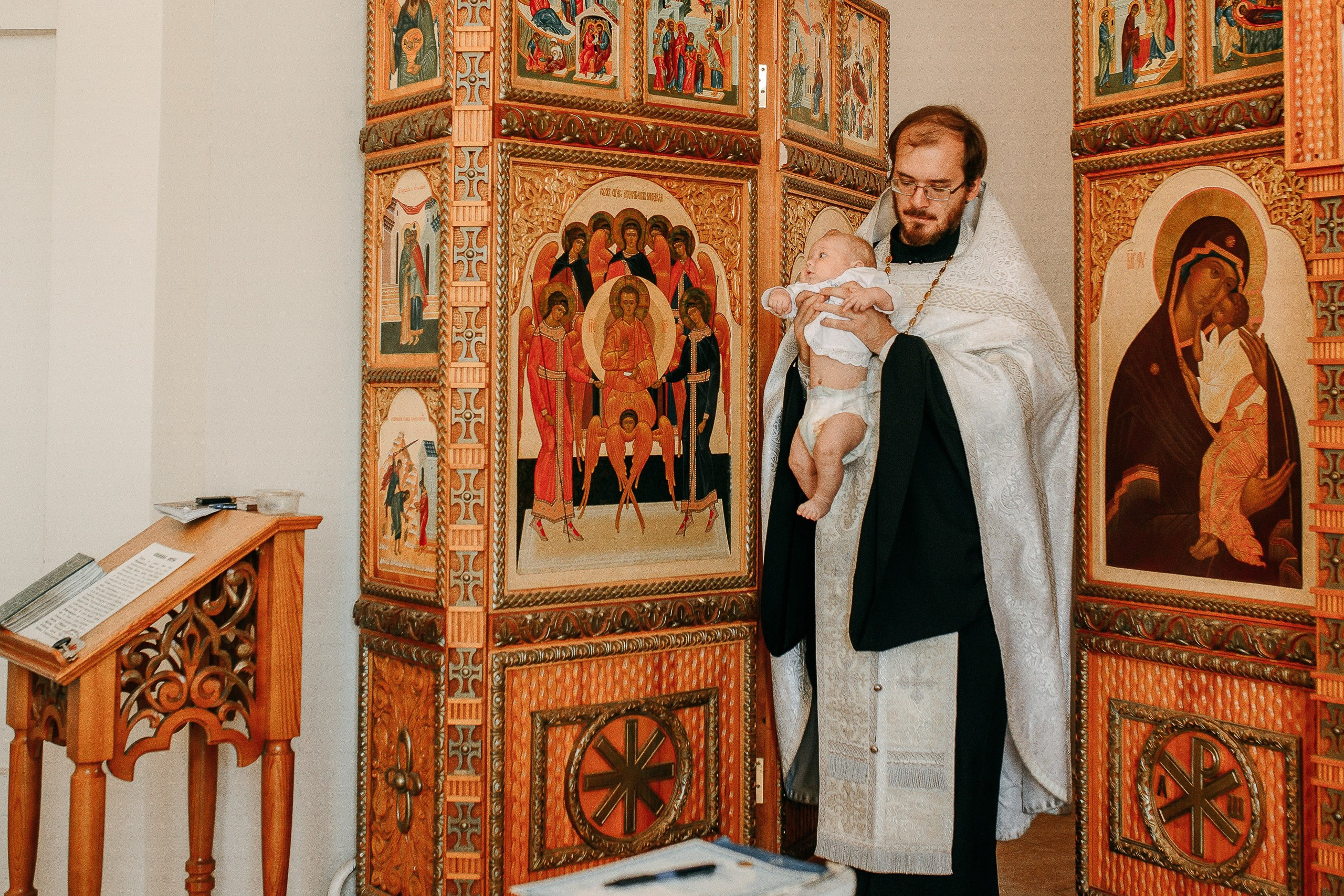 Профессиональная съемка таинства крещения: священник держит младенца на руках на фоне иконостаса.