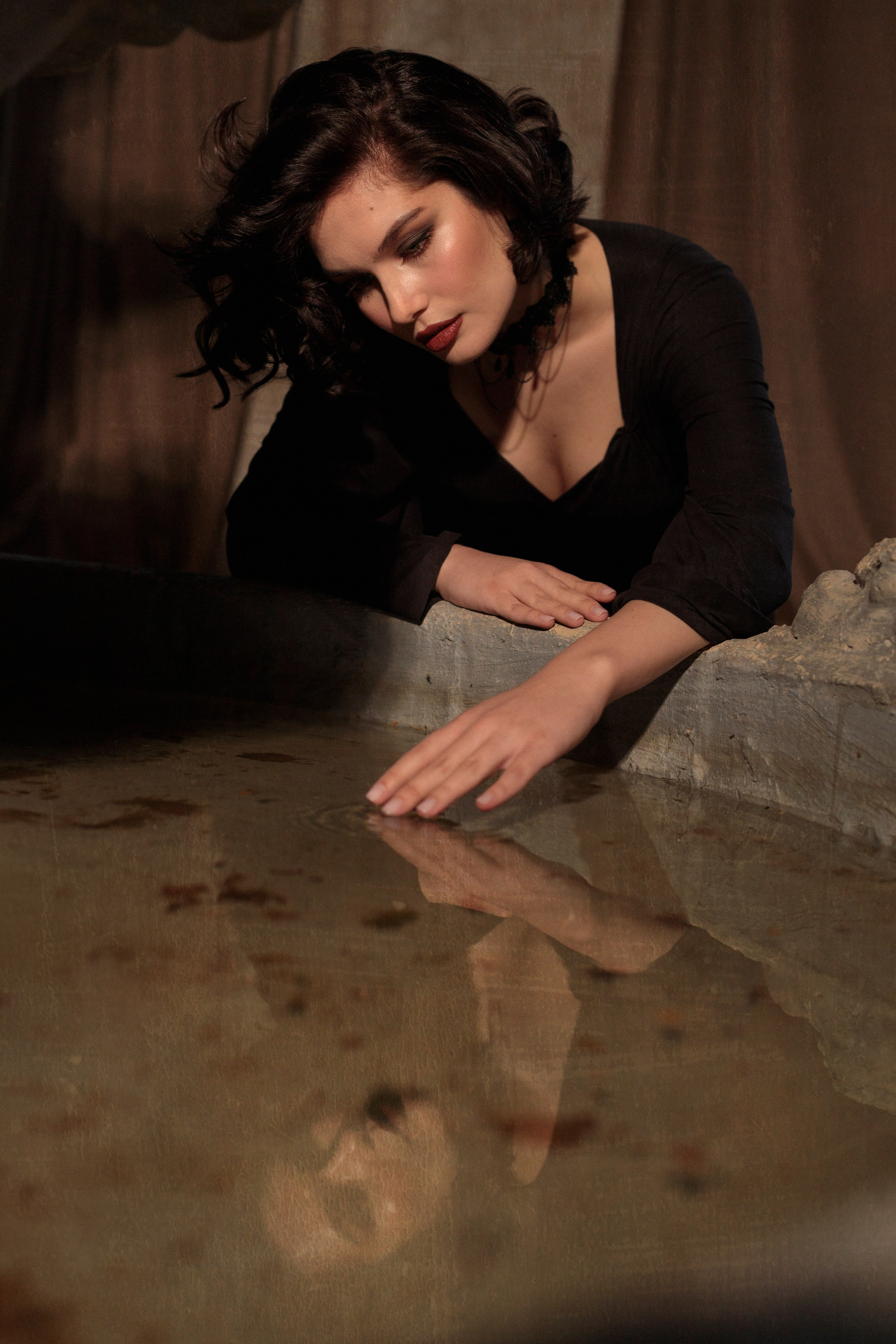 Девушка склонилась над фонтаном, рукой касается воды, в воде видно отражение девушки