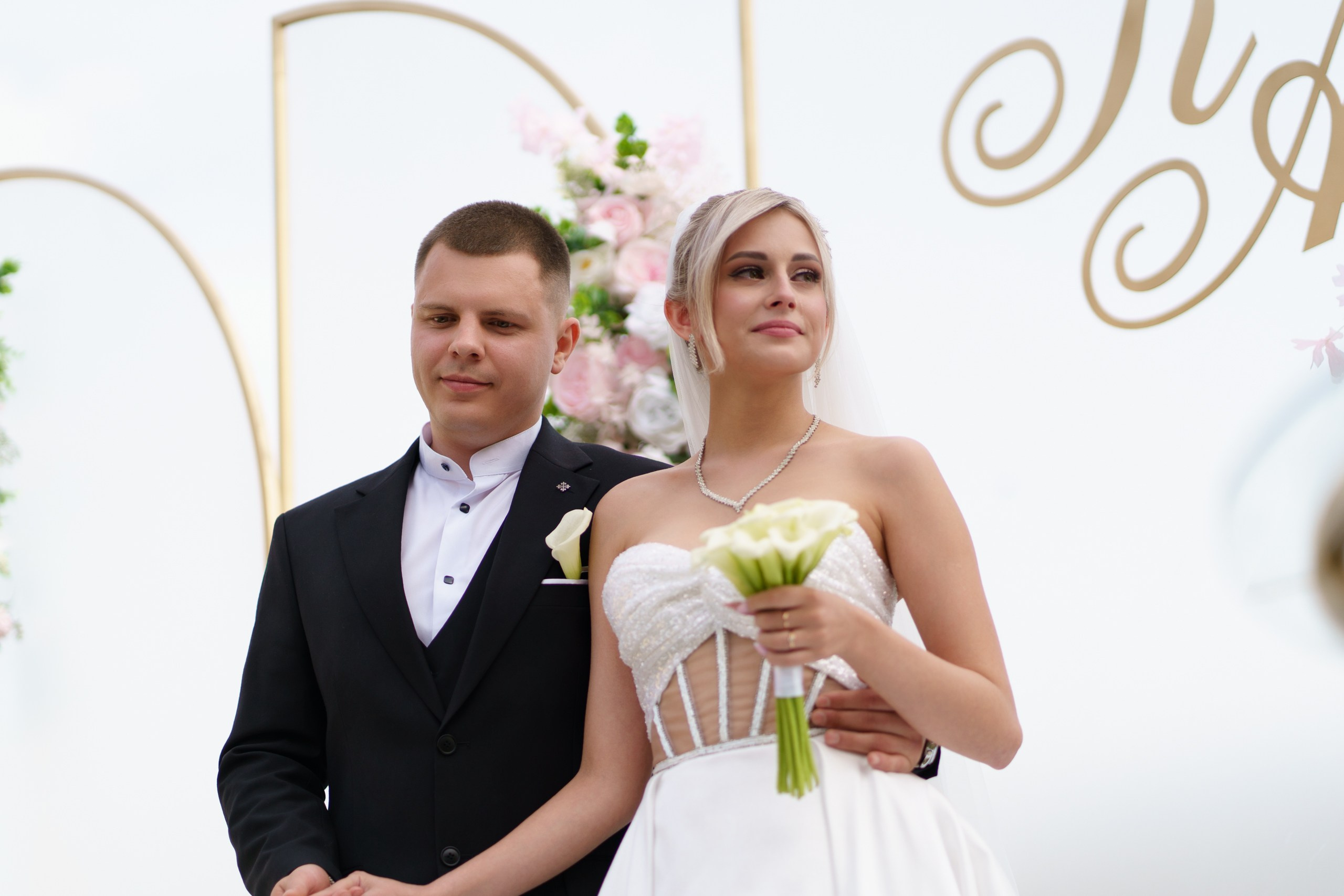 Романтическая сцена бракосочетания: жених в классическом чёрном костюме и невеста в роскошном белом платье стоят рядом. Невеста держит букет калл, а позади них — праздничная арка с золотыми деталями | фотограф на торжественную регистрацию Екатеринбург