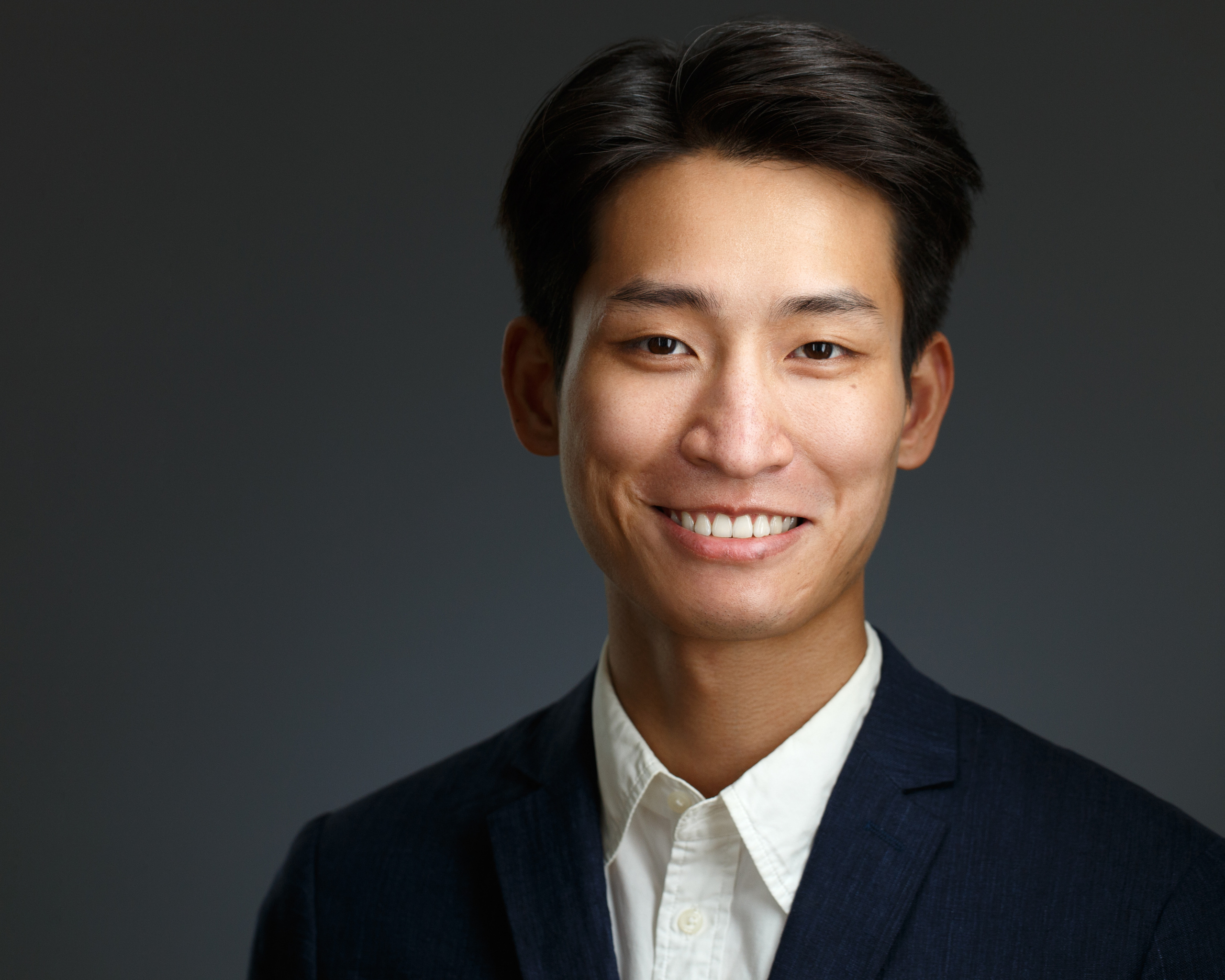 Хедшот. Бизнес-портрет молодого улыбающегося китайца в тёмном пиджаке и белой рубашке без галстука на тёмном фоне.