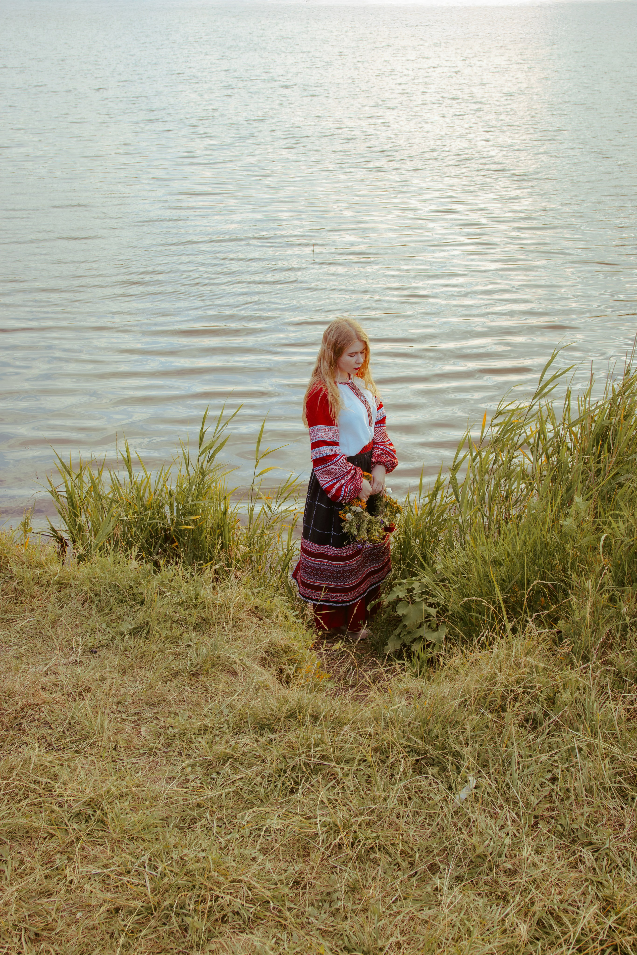 Грустная девушка стоит возле реки с венком из полевых цветов в руках