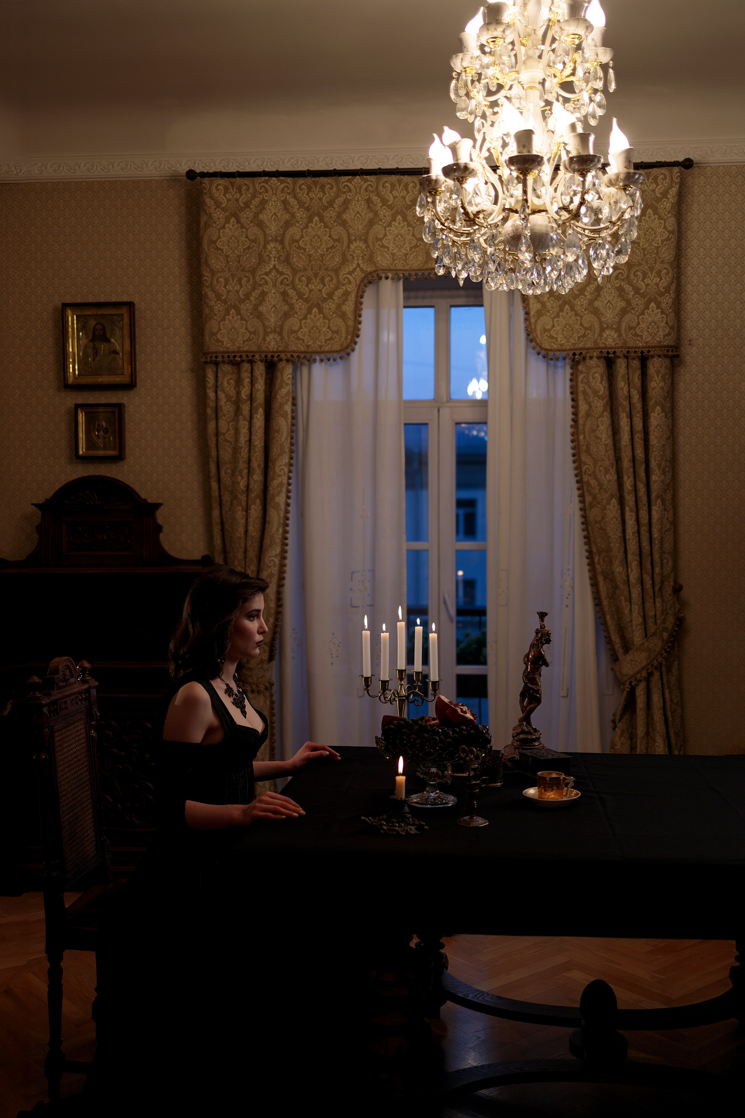 Дальний план, где девушка сидит за столом, над головой светит яркая винтажная люстра, на столе стоит канделябр с горящими свечами.