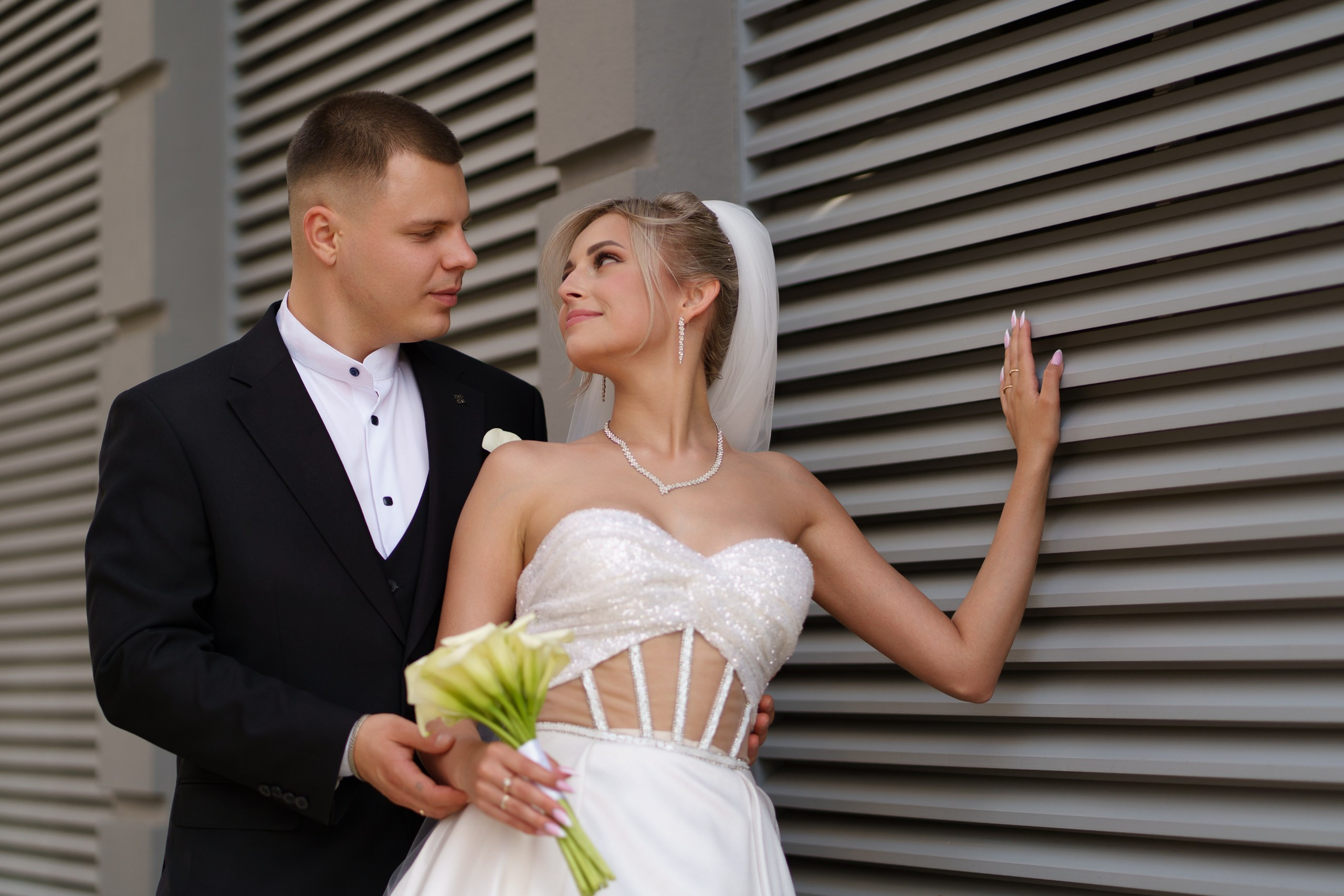 Невеста в роскошном белом платье с открытыми плечами и фатой и жених в элегантном чёрном костюме запечатлены в динамичной позе: жених держит невесту, приподнимая её ногу. Они стоят на фоне современного здания | портреты на свадьбе Екатеринбург